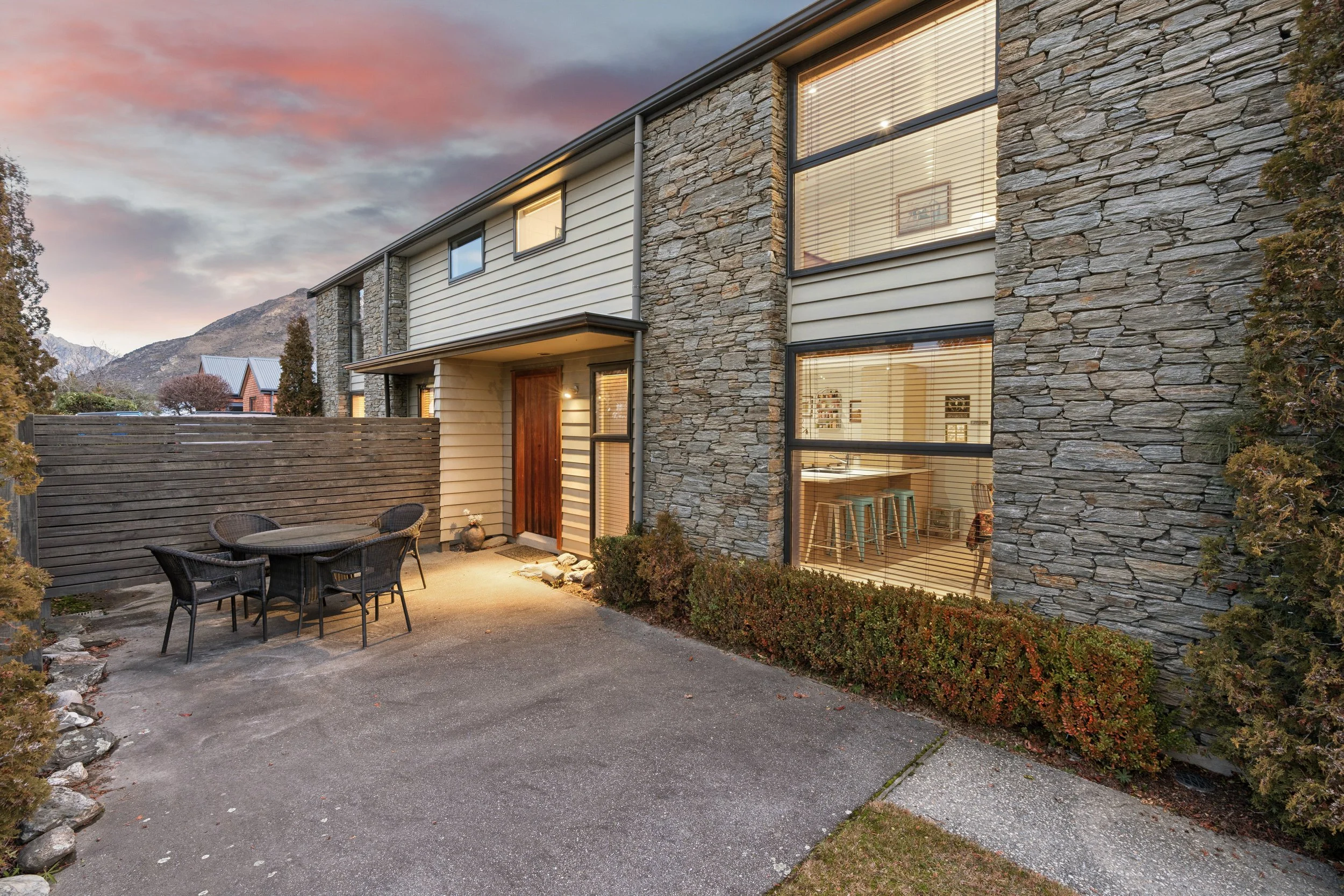 A modern house with large windows and a stone facade, view of a backyard patio with outdoor seating, and a sunset sky in the background.