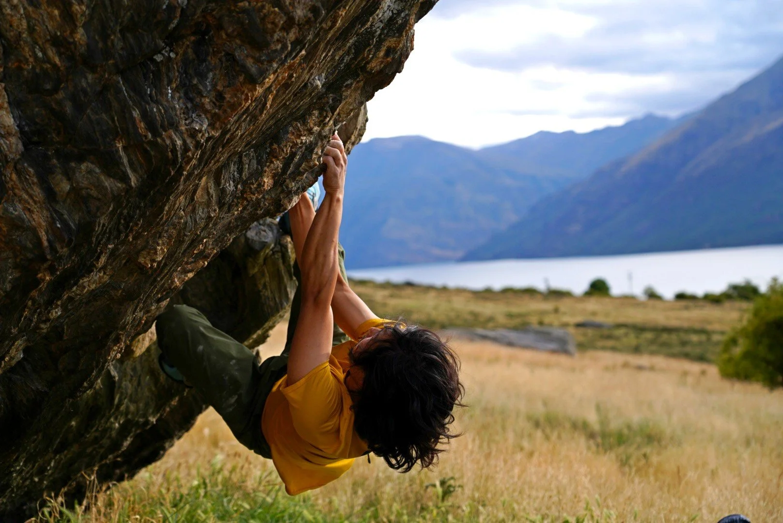 A person rock climbing on an overhanging rock face in an outdoor natural setting with mountains, a lake, and open fields in the background.