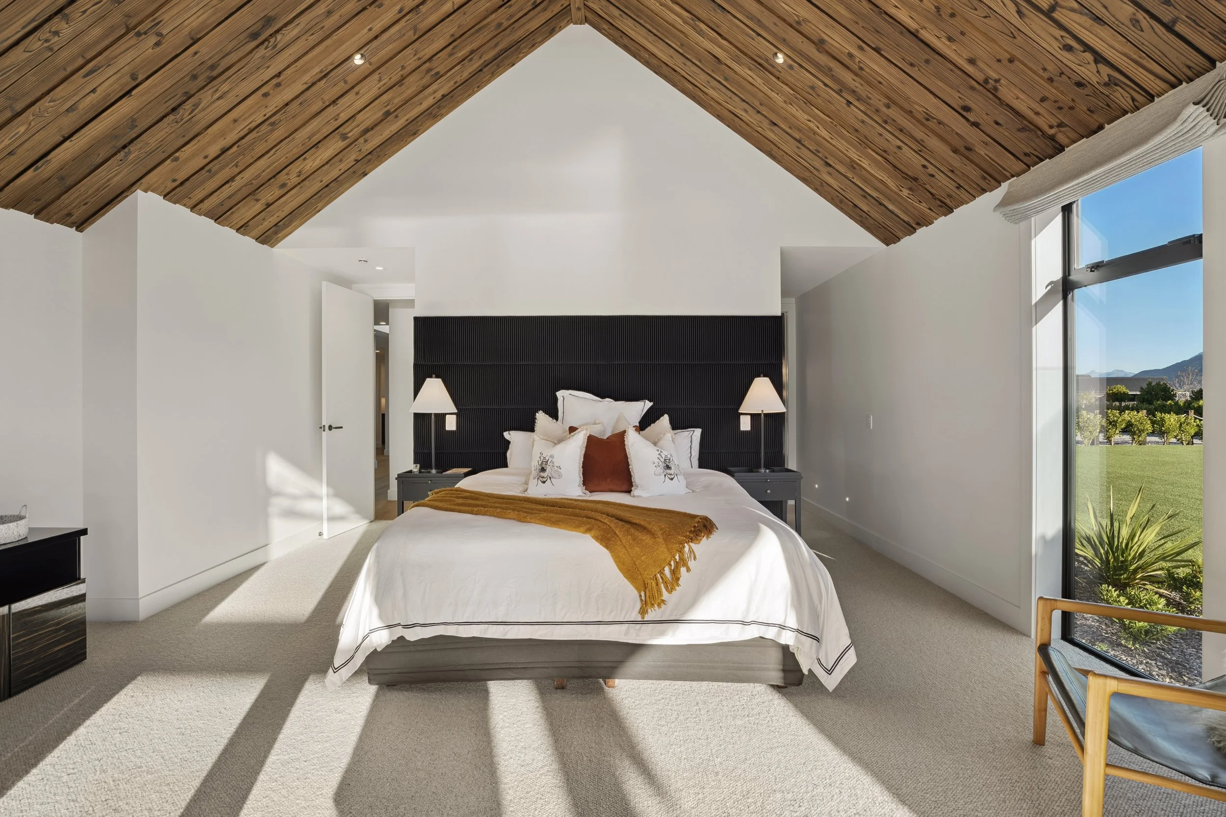 Bright bedroom with high wooden vaulted ceiling, large window showing a landscape, white walls, a bed with white bedding, black headboard, nightstands with lamps, and a small sitting area with a wooden chair.