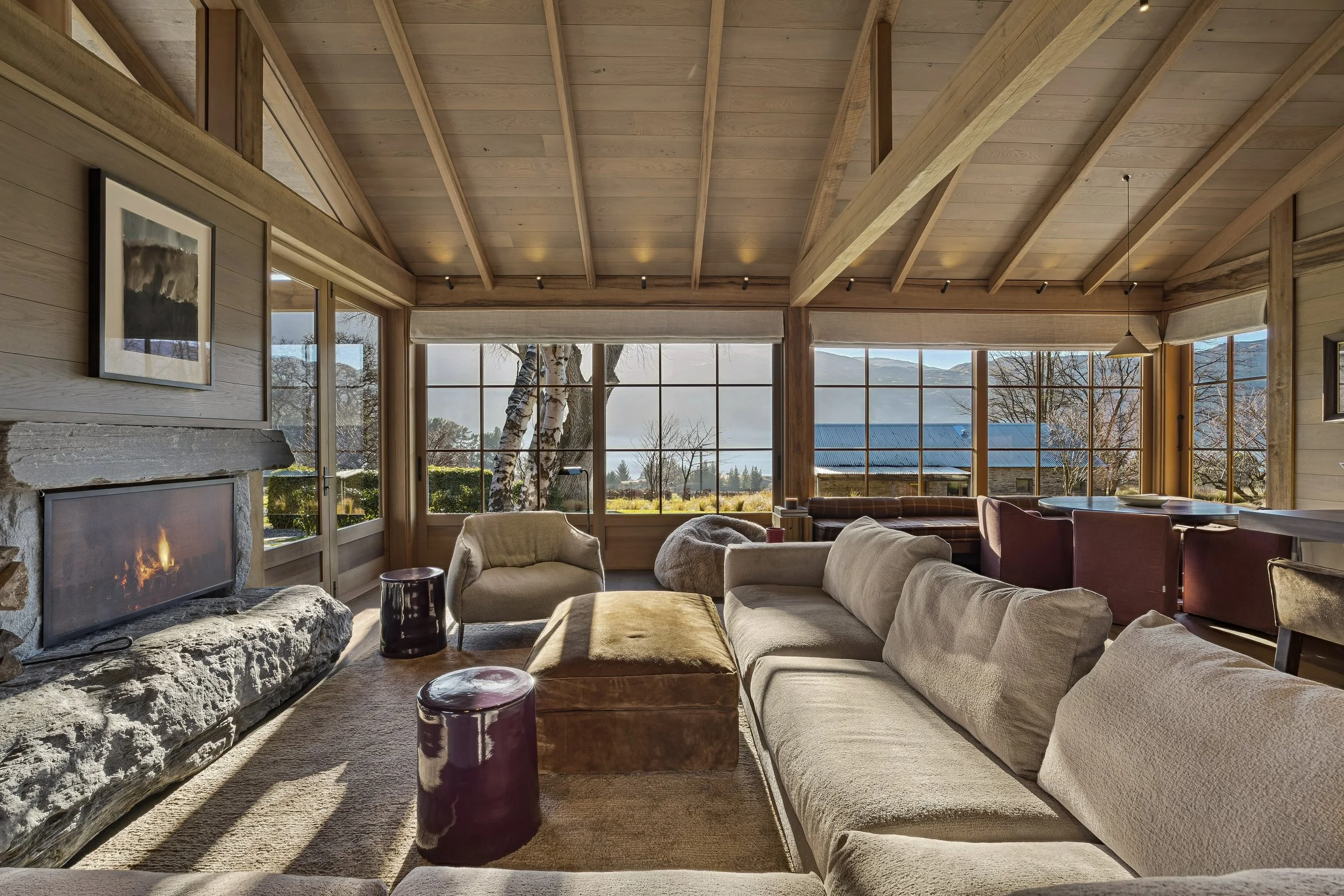 Interior of a cozy living room with large windows showing a scenic mountain view, wooden ceiling beams, a stone fireplace with a fire burning, and multiple sofas and chairs arranged for relaxation.