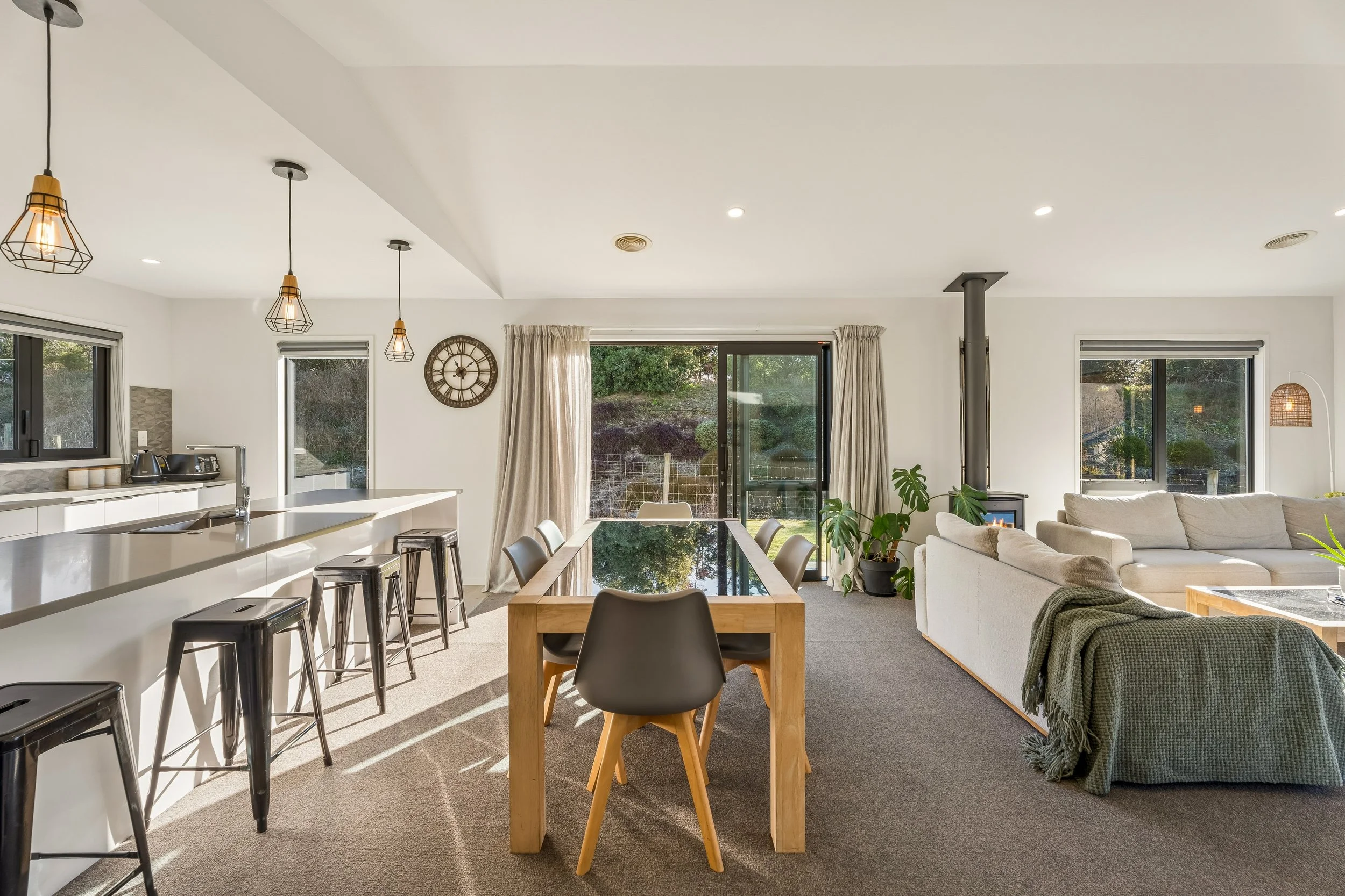 Open concept living area with a kitchen, dining table, and seating area, featuring large windows, pendant lights, and a wall clock.