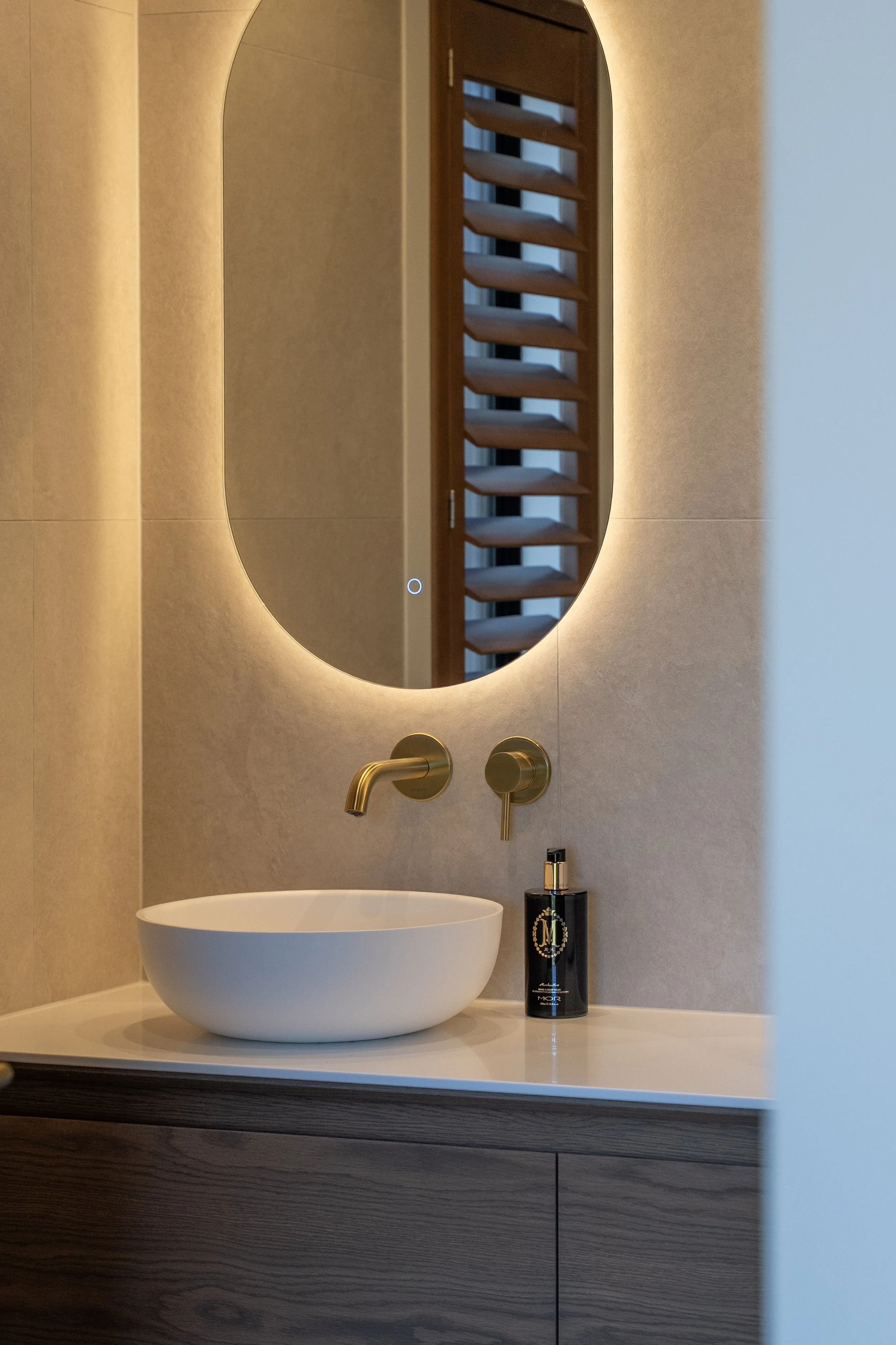 A modern bathroom with a round vessel sink, a wall-mounted faucet, a black bottle of soap, a large oval mirror with backlighting, and a wooden slatted window or decorative panel behind the mirror.