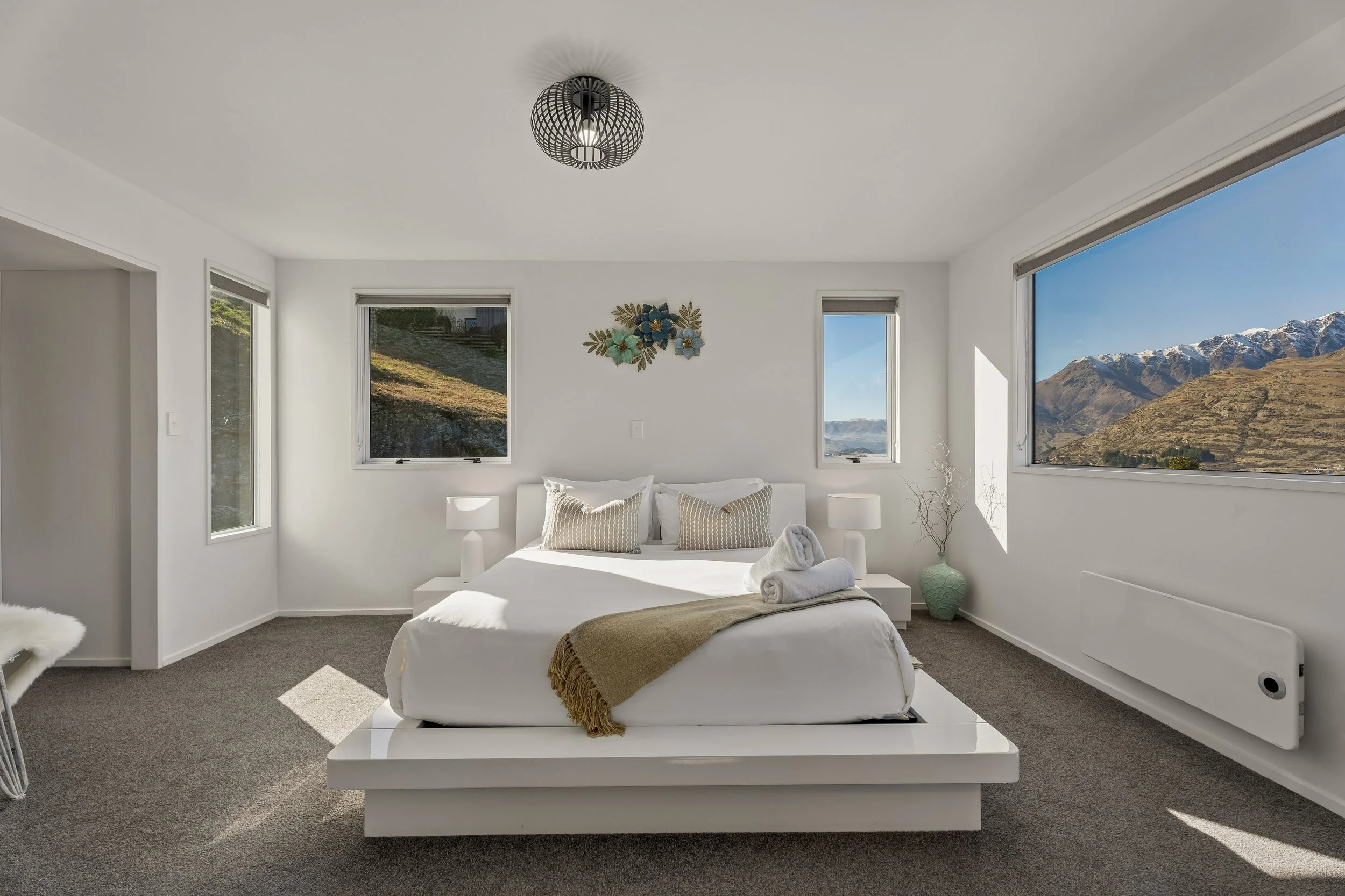 Bright bedroom with white walls, large window views of mountains and blue sky, white bed with beige blanket and rolled white towels, decorative wall art, two bedside lamps, gray carpet, modern chandelier.
