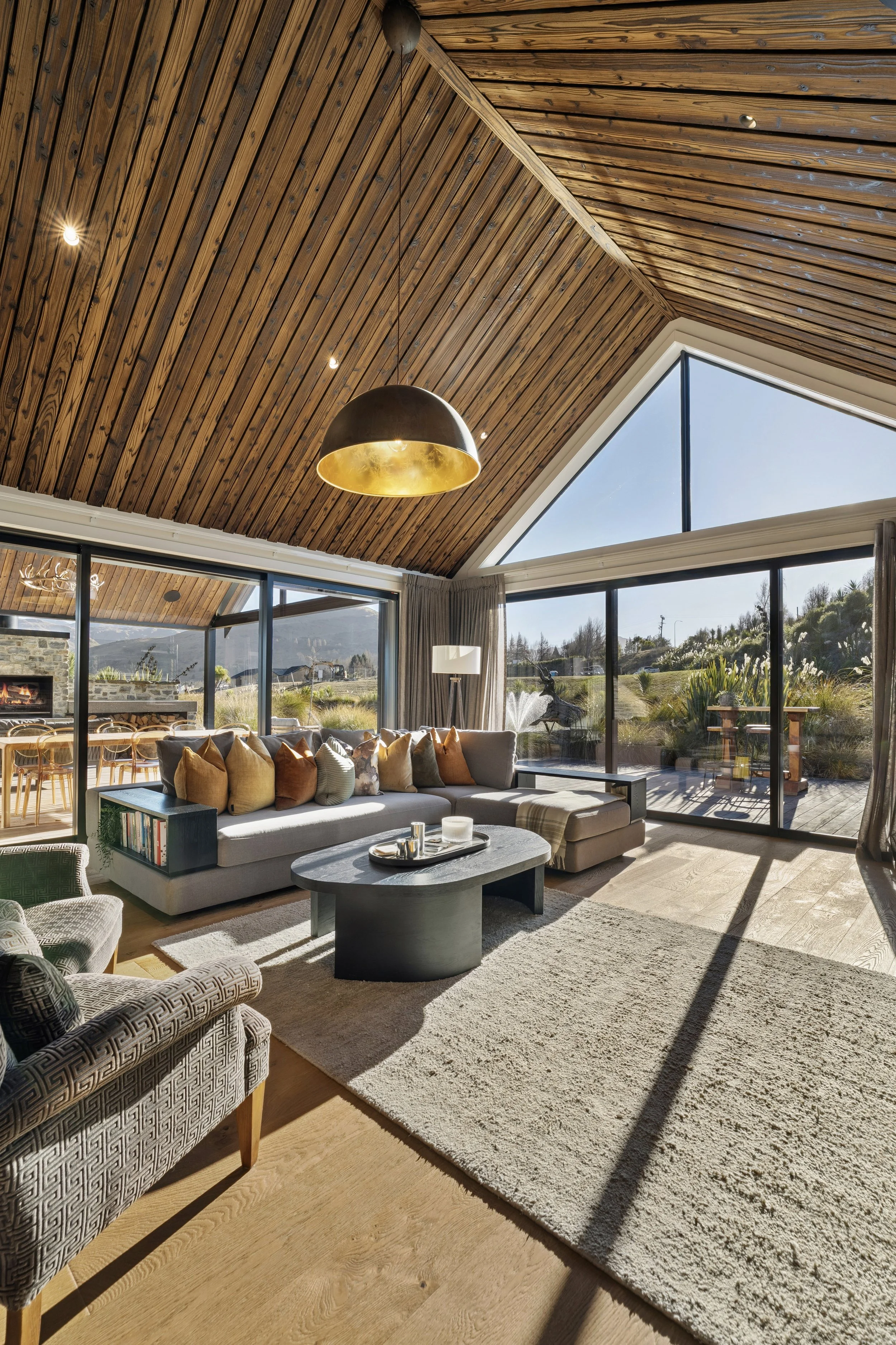 Living room with vaulted wooden ceiling, large windows, beige sofa with colorful pillows, round coffee table, patterned armchair, and a cream rug. Outside view shows a garden with a fountain and trees.