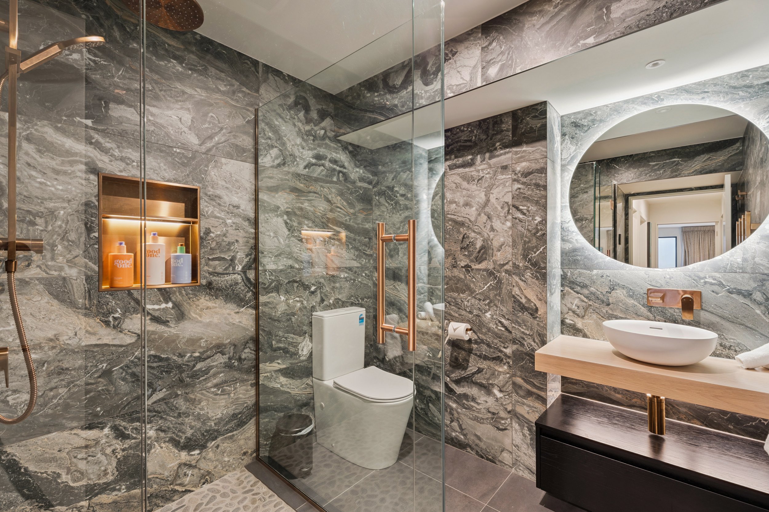 Modern bathroom with gray marble walls, a glass shower enclosure, a wall-mounted mirror with backlighting, a white vessel sink on a wooden vanity, and accessories like soap bottles and a towel.