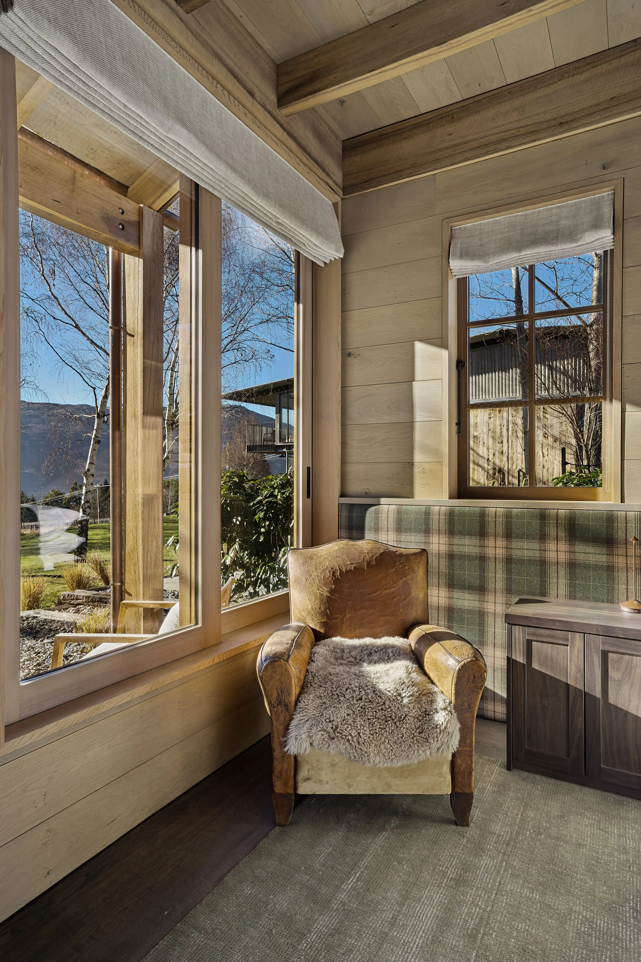Interior of a room with wooden walls and large windows showing trees and mountains outside. There is a vintage armchair with a fur throw and a plaid upholstered bench next to a small side table.