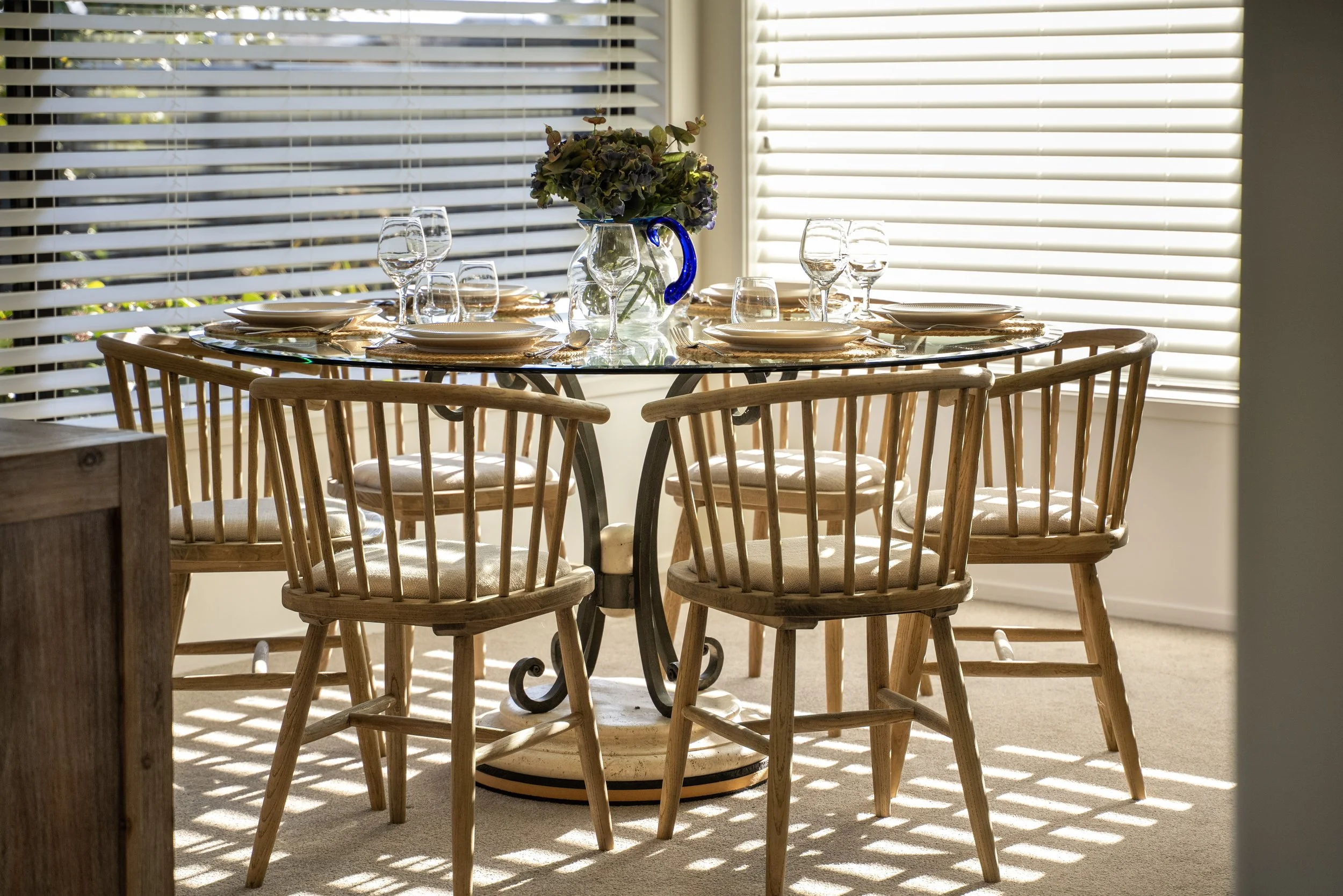 A round dining table set with plates, glasses, and cutlery in front of sunlit window blinds, with a large floral arrangement in a glass vase as a centerpiece.