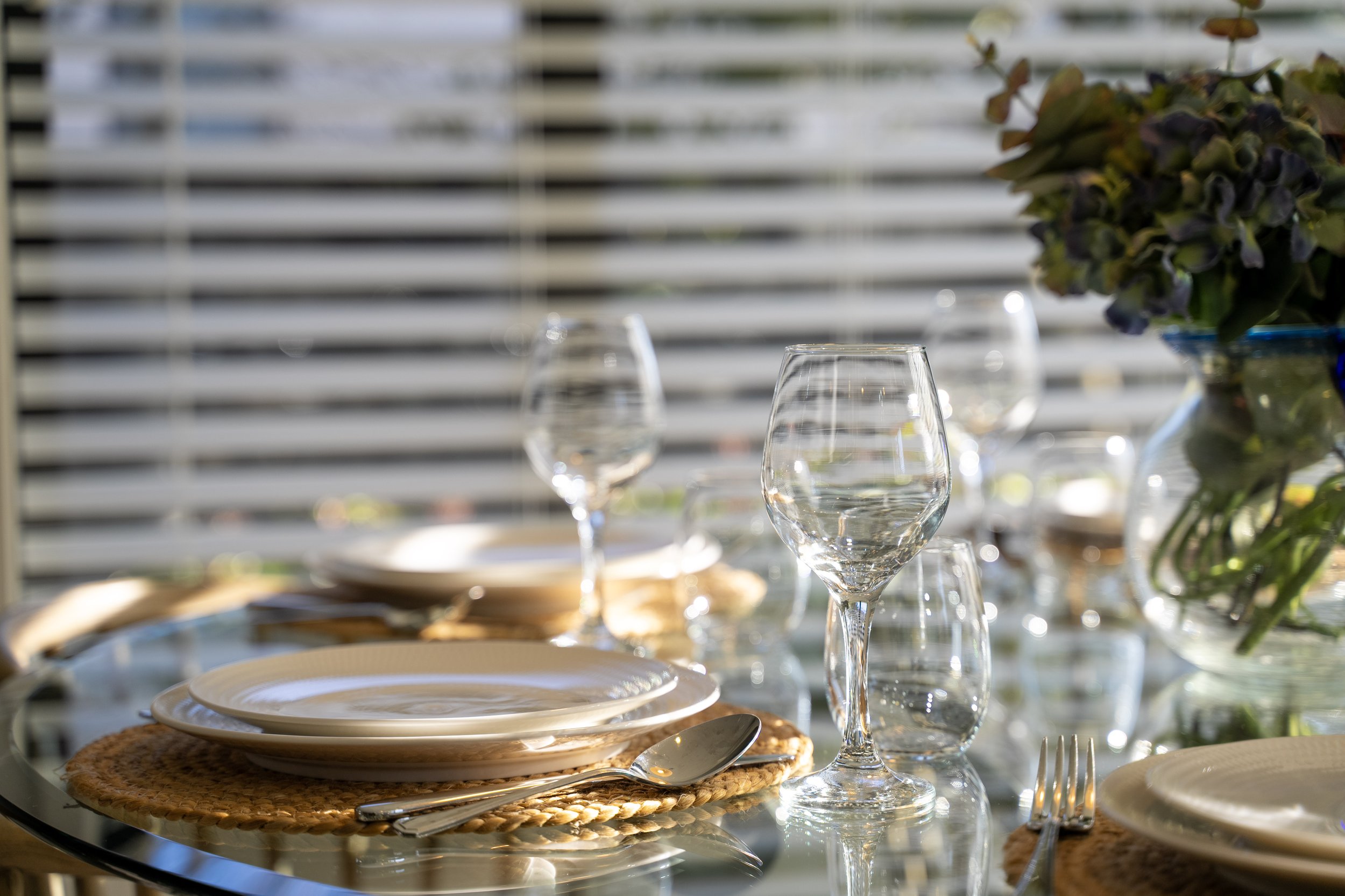 A dining table set with plates, silverware, and empty wine glasses, with a backdrop of horizontal window blinds and a flower arrangement on the right.