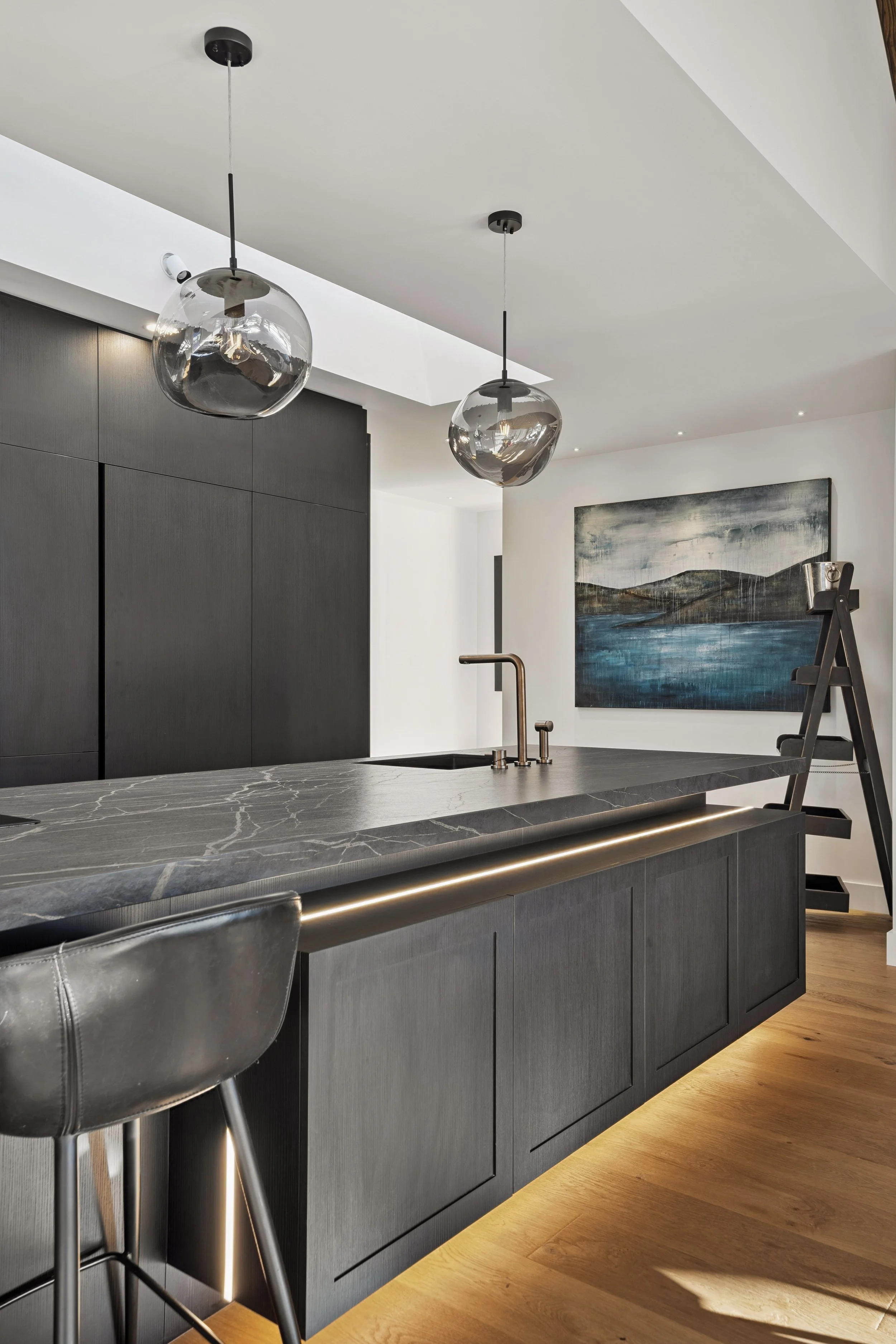 Modern kitchen with dark cabinetry, a marble countertop, two glass pendant lights, a painting on the wall, and warm under-cabinet lighting.