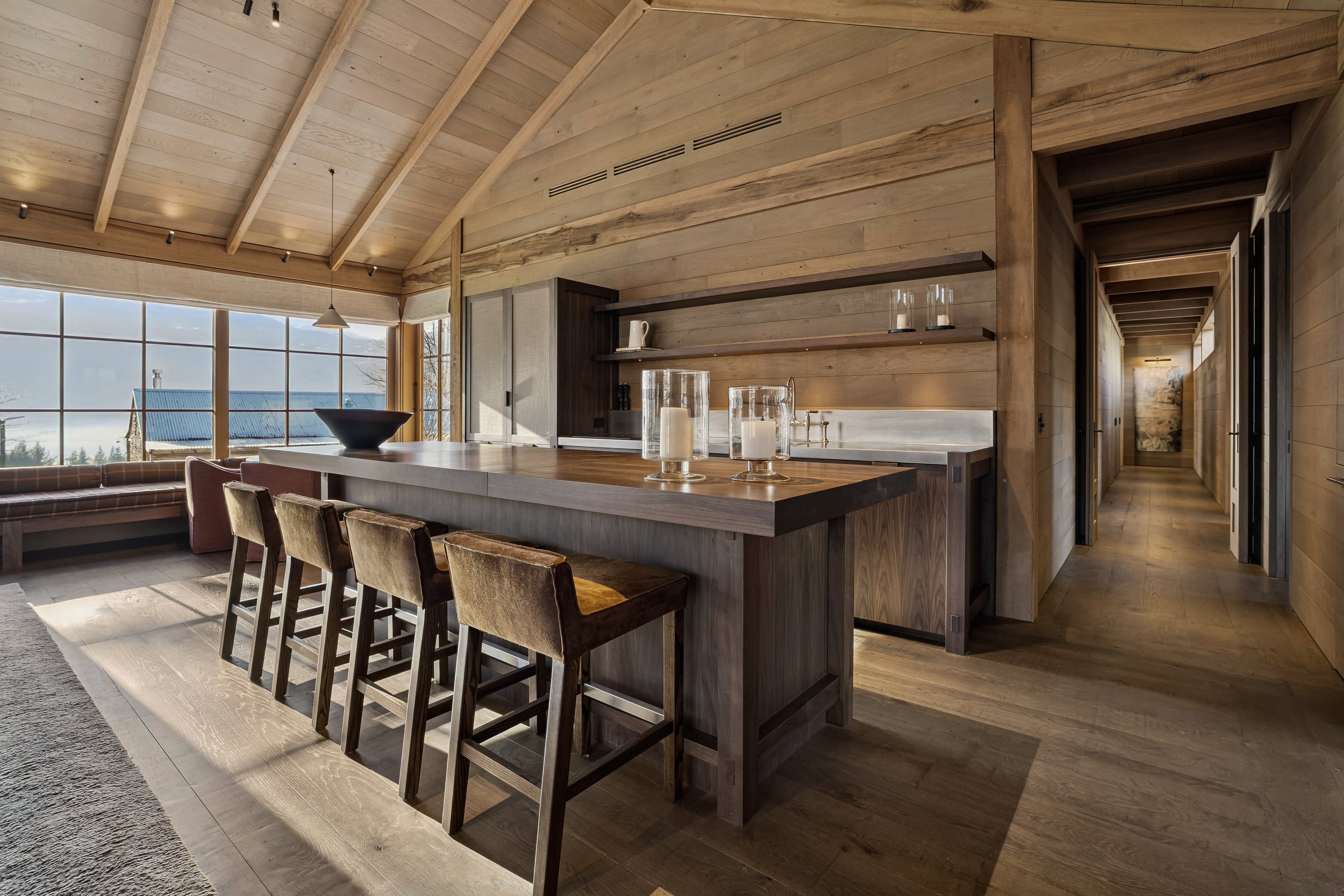 Interior view of a wooden kitchen and dining area with a high, vaulted ceiling, large windows offering a scenic outdoor view, a kitchen island with barstools, and decorative candles on the counter.