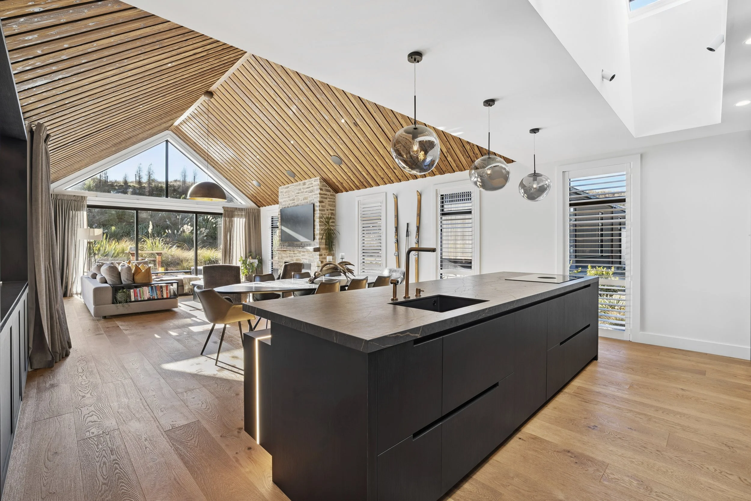 Modern kitchen and living room with vaulted ceiling, wooden accents, large windows, and sleek black island.