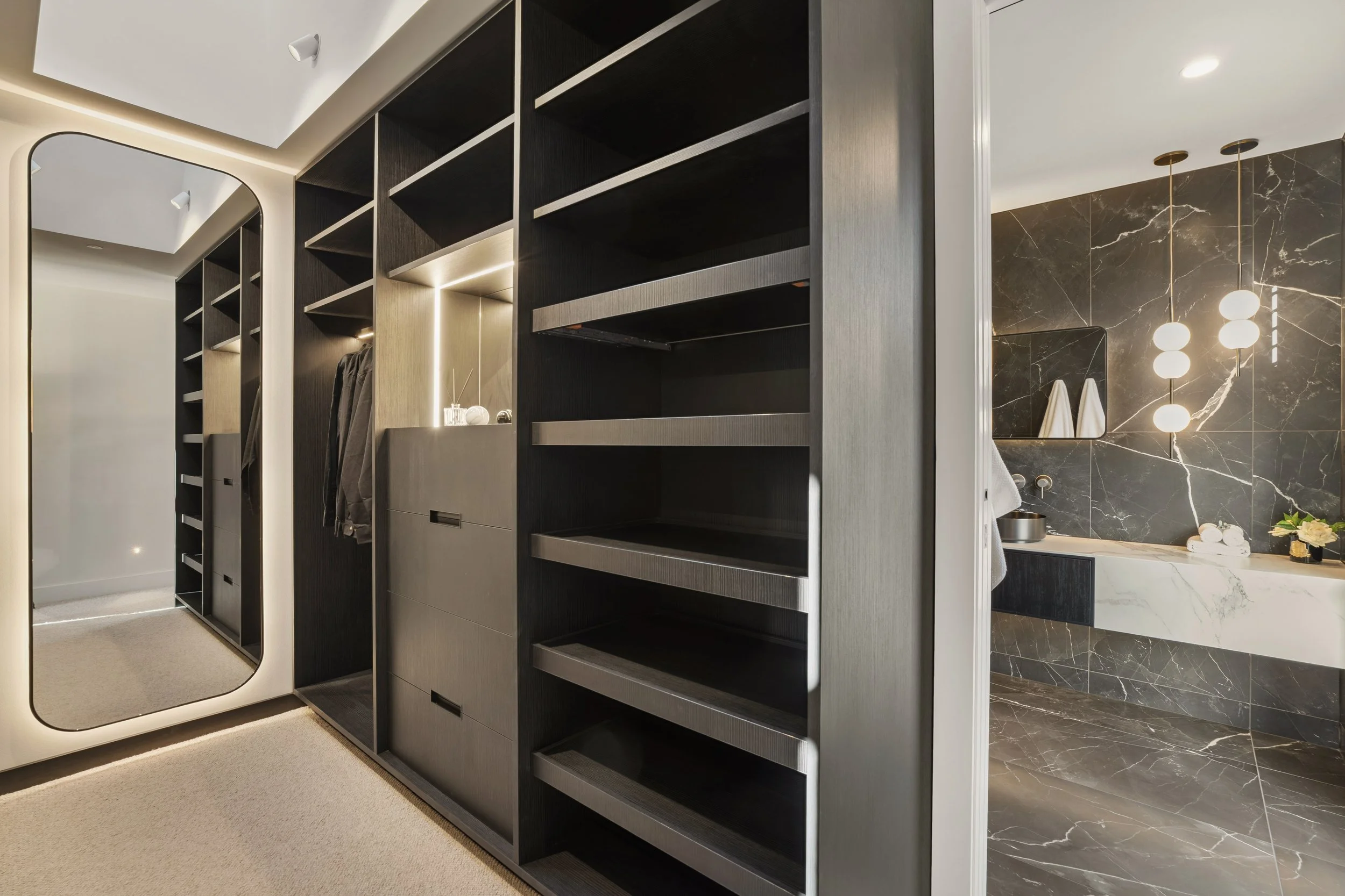 A modern walk-in closet with black shelving and a full-length mirror. The closet is adjacent to a bathroom area with a dark marble wall, pendant lights, and a vanity with decorative items.