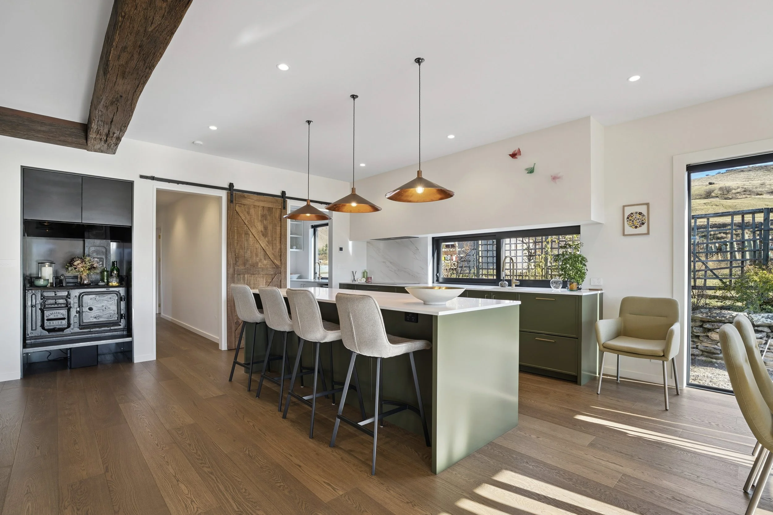 Open-concept kitchen with white walls, wooden floor, and modern decor. Features a kitchen island with beige chairs, green cabinetry, and pendant lighting. Large window shows a scenic outdoor landscape.