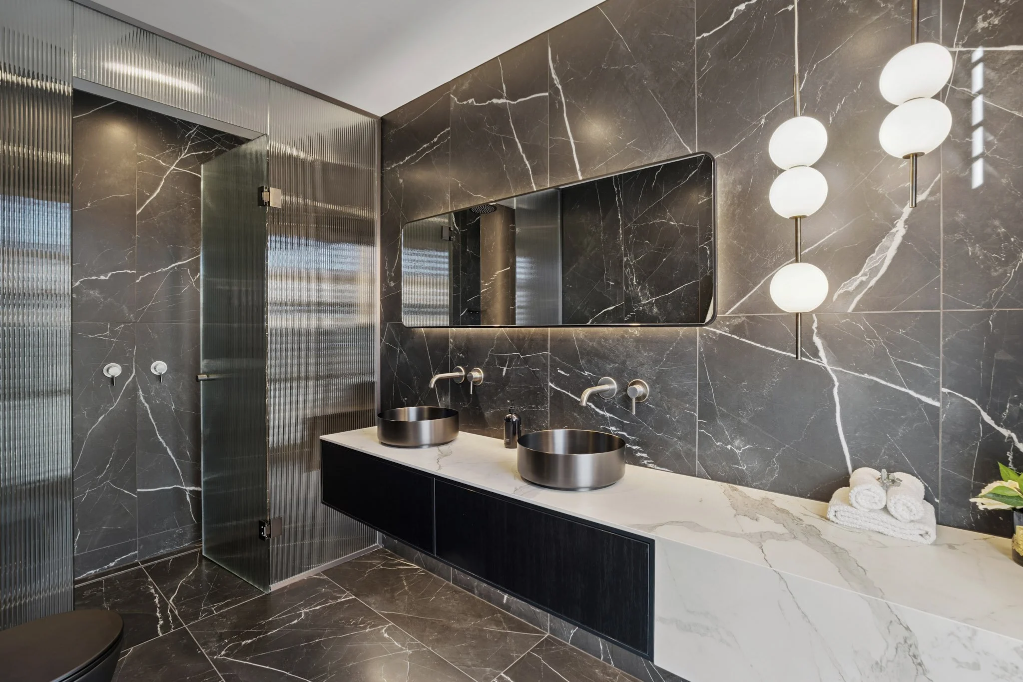 Modern bathroom with black and white marble walls and floor, double sinks with round stainless steel basins on a white marble countertop, large mirror, spherical white pendant light fixture, green plants, and neatly folded white towels.