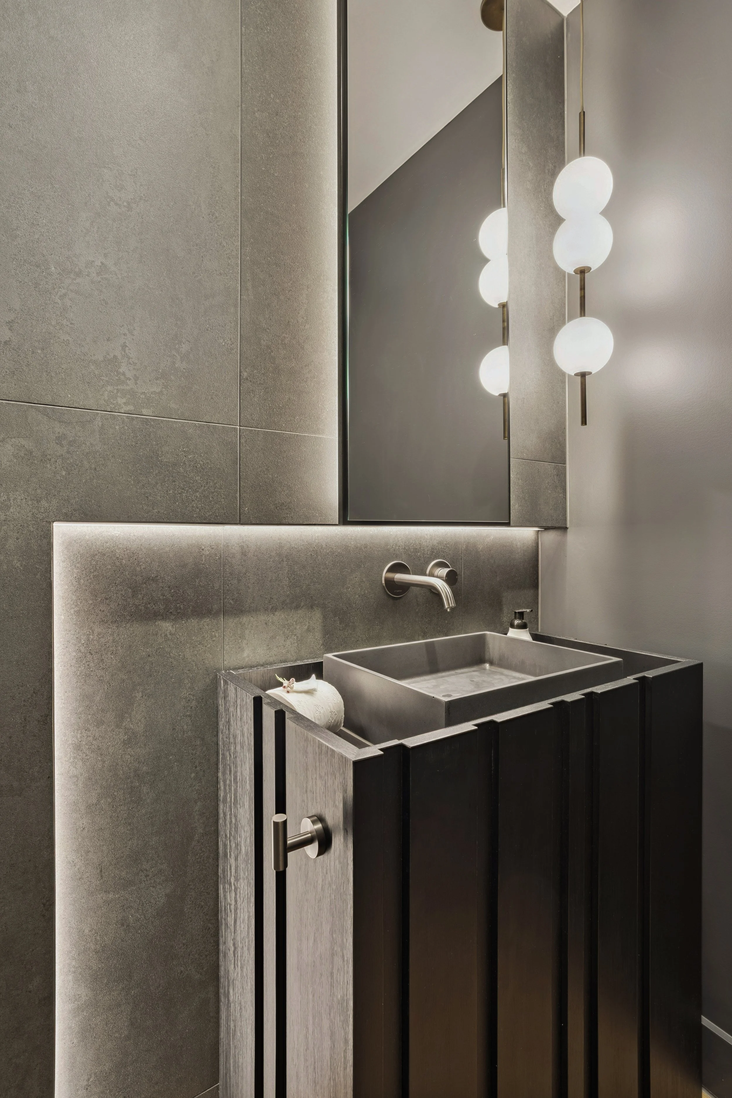 A modern bathroom with a gray textured wall, a wall-mounted faucet, a black sink with a rolled towel and flowers, a large mirror, and a vertical strip of three spherical light fixtures.