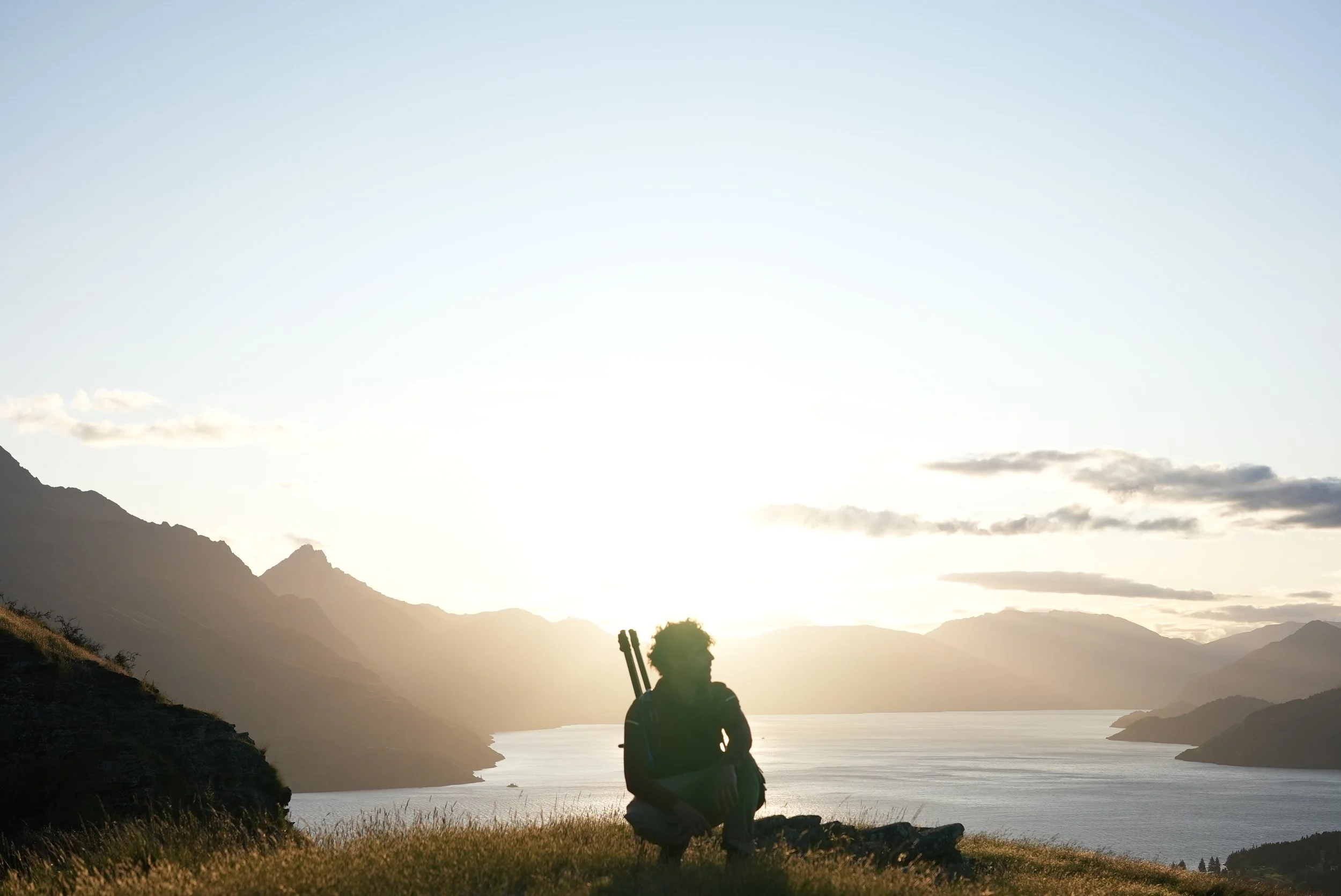 Person sitting on a grassy hill with mountains and a water body at sunset in the background