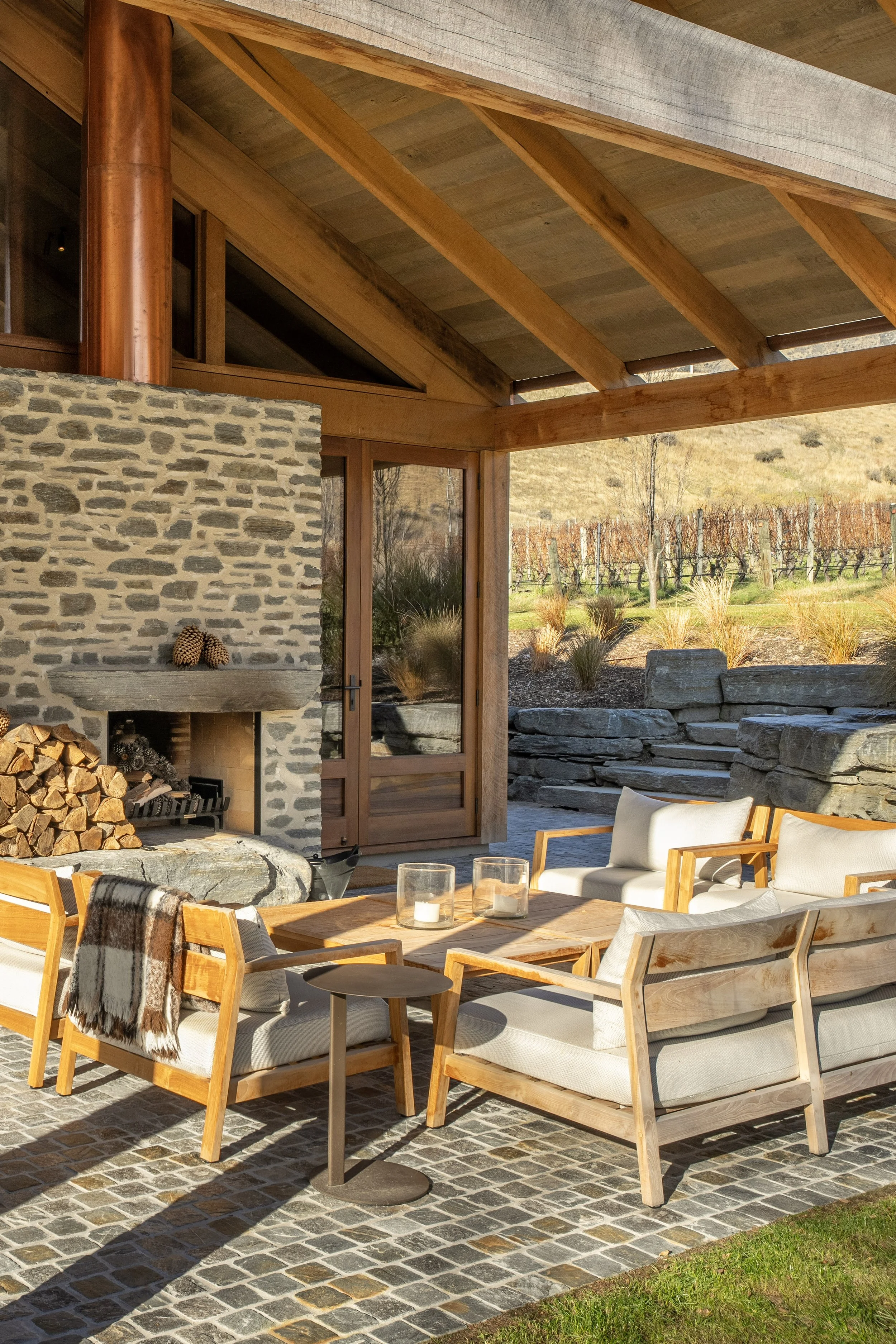 Outdoor patio area with stone fireplace, wooden furniture, cushioned chairs, and a table with candles, overlooking a vineyard in a rural landscape.