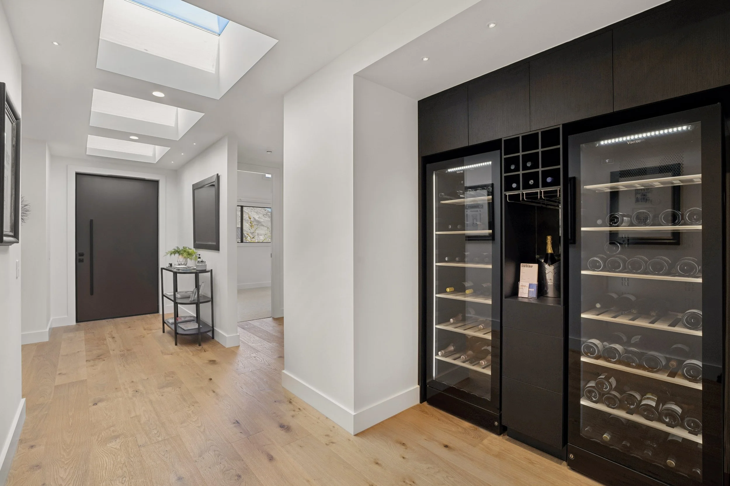 A modern interior with white walls and light wooden flooring. Features a black wine cooler stocked with wine bottles, a small black table with plants and books, and ceiling skylights providing natural light.
