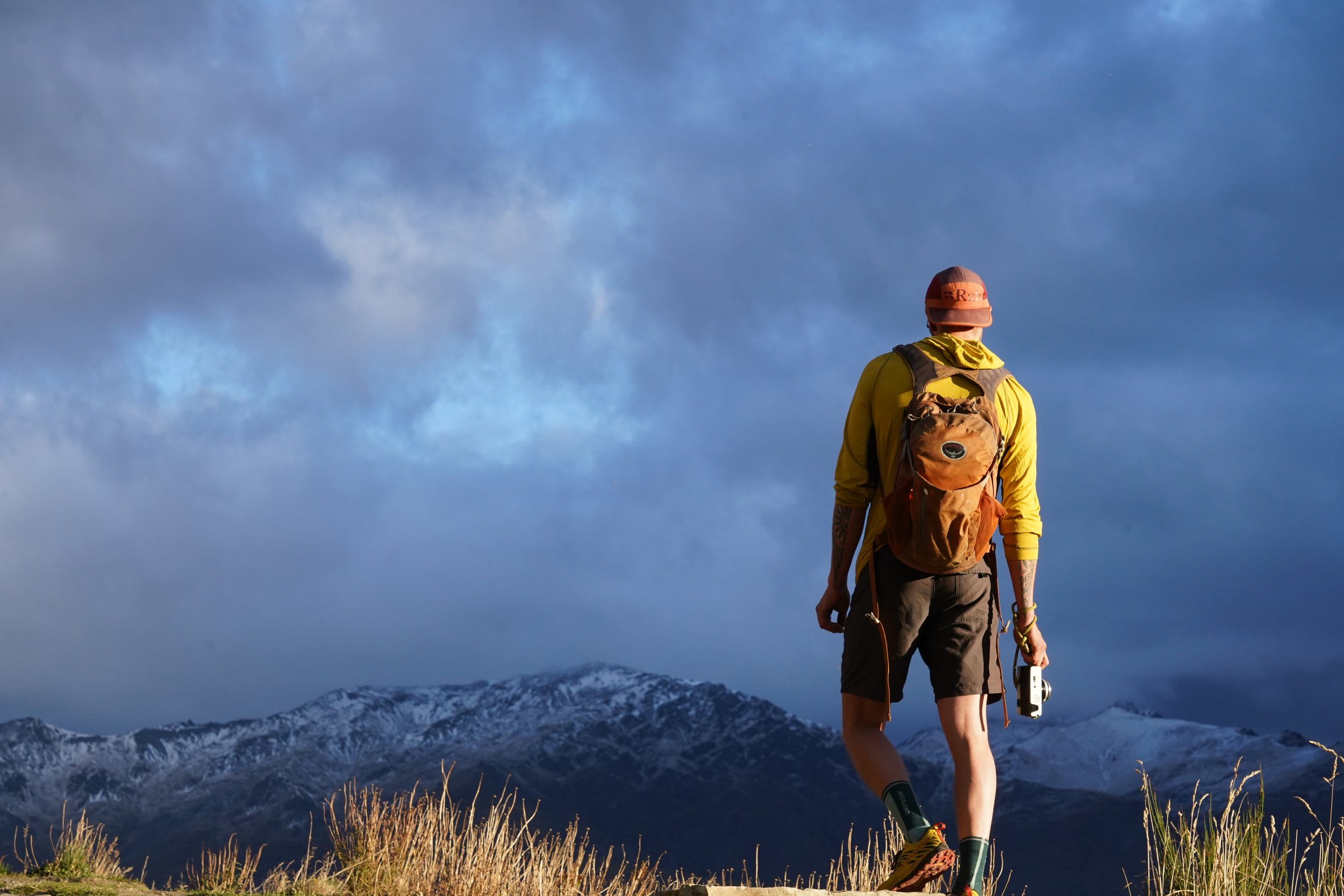 A hiker dressed in yellow with a backpack and hat, holding a walking stick and a GPS device, walking in a mountainous area with snow-capped peaks and a partly cloudy sky.
