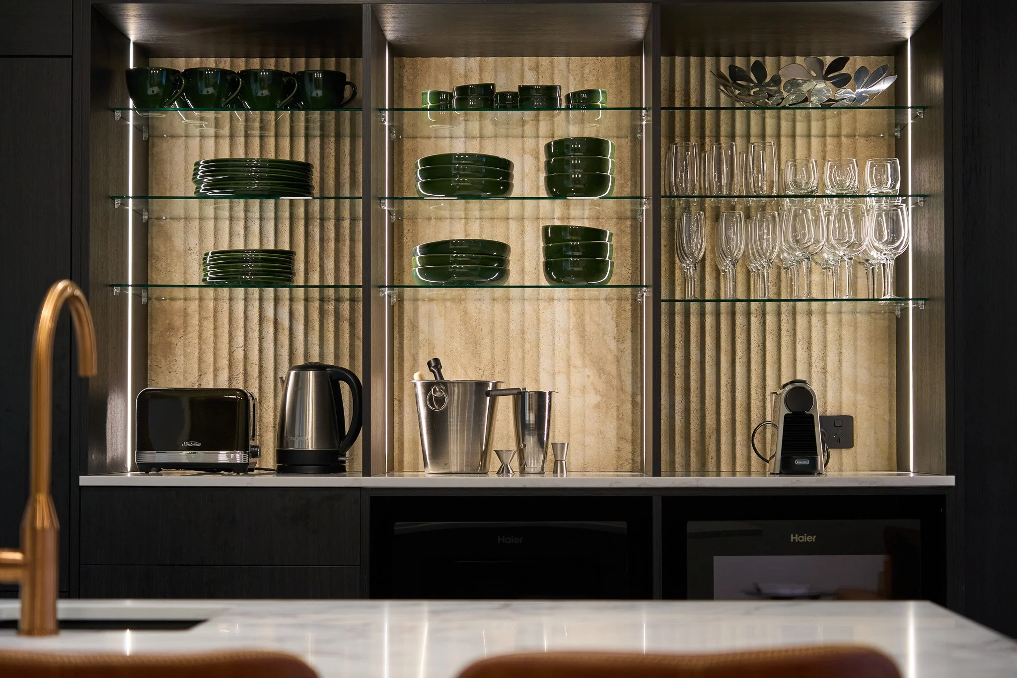 Kitchen cabinet with glass shelves holding dark green cups, bowls, and a decorative metal bowl. Below are appliances and utensils including a toaster, electric kettle, mixing bowl, and glasses.