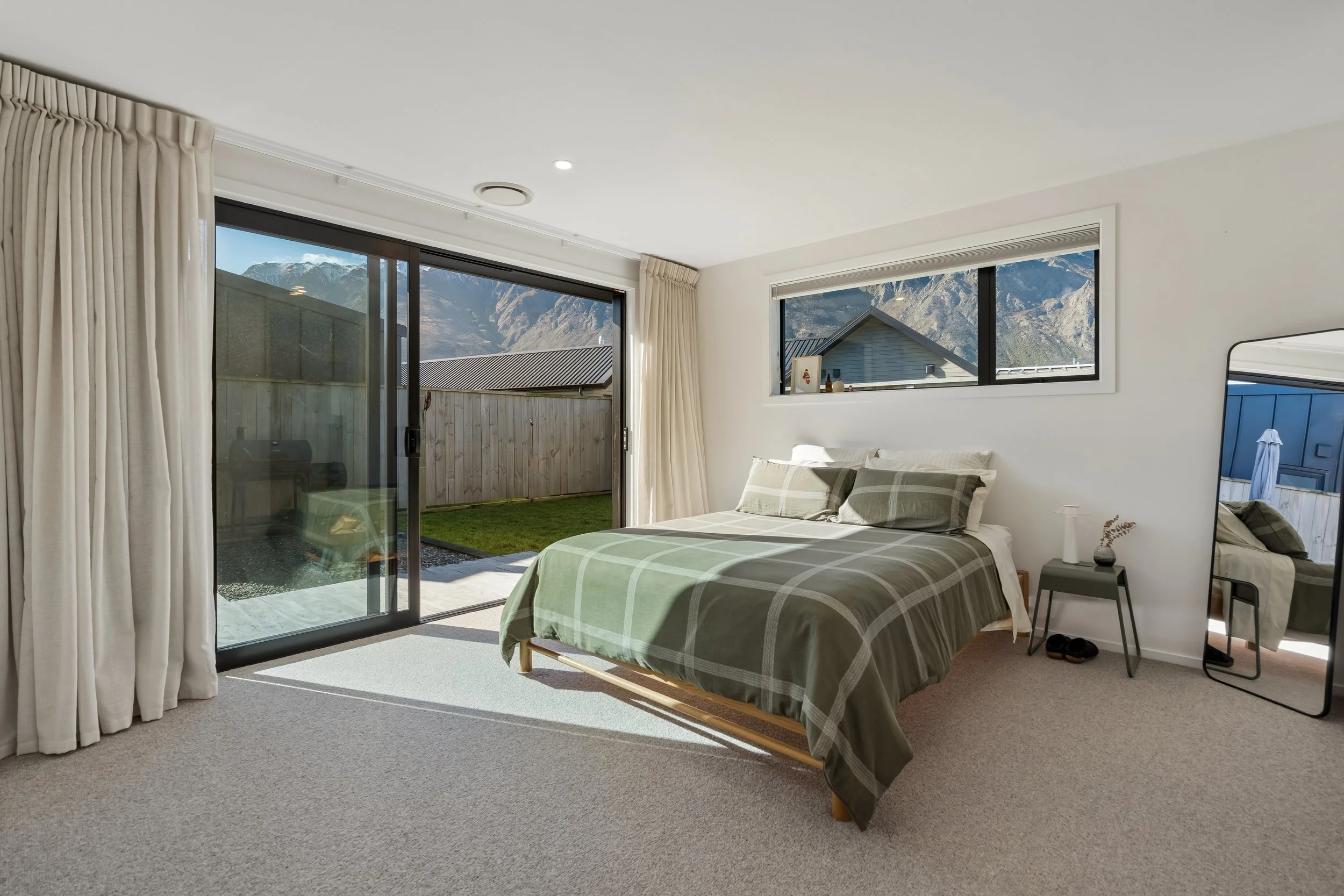 A bedroom with a bed covered in green plaid bedding, a bedside table with a small lamp and vases, large sliding glass door opening to a backyard, and a window showing mountains in the background.
