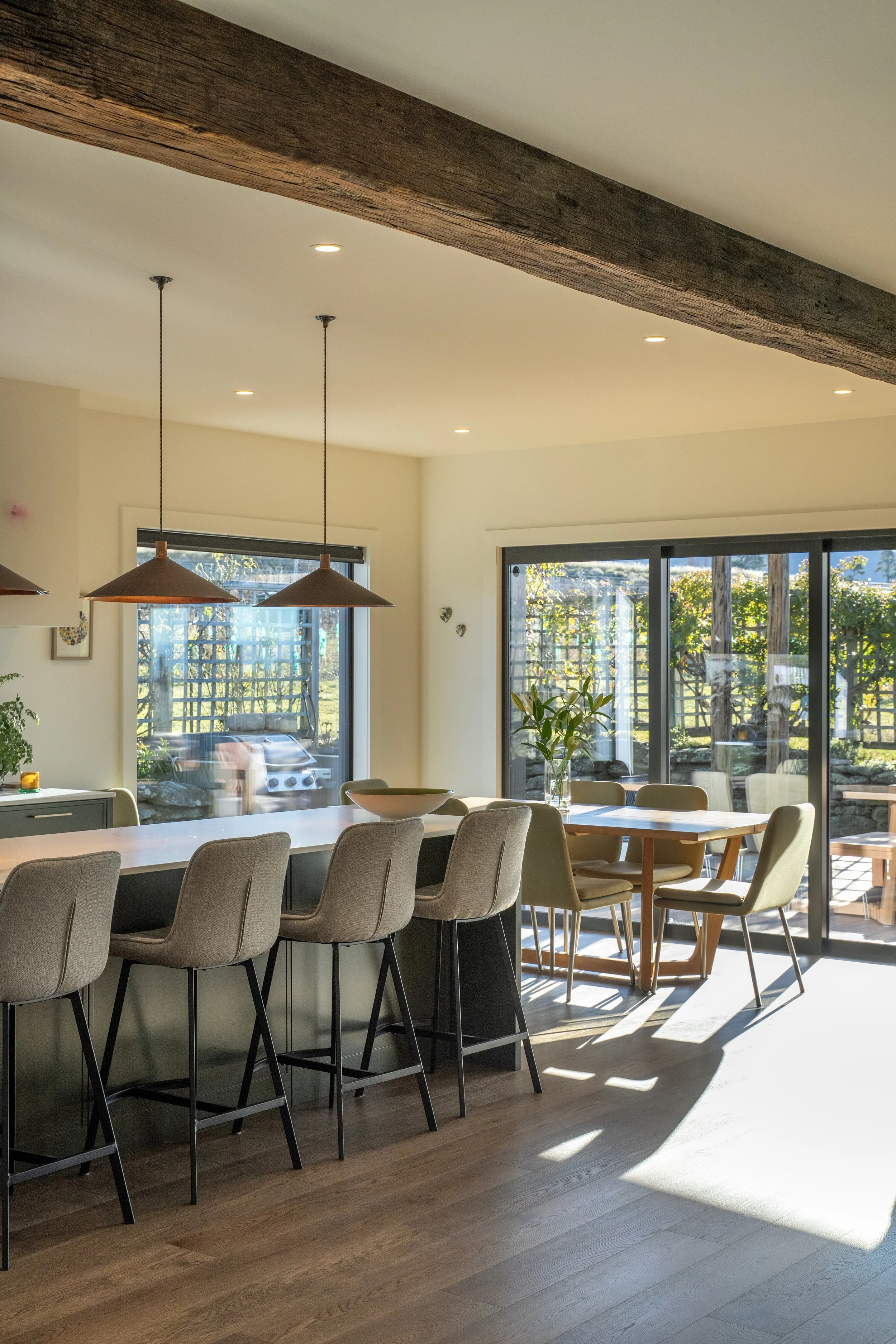 A modern dining area with a large white table surrounded by beige upholstered chairs, natural sunlight coming through large sliding glass doors, and two brown pendant lights hanging from the ceiling.