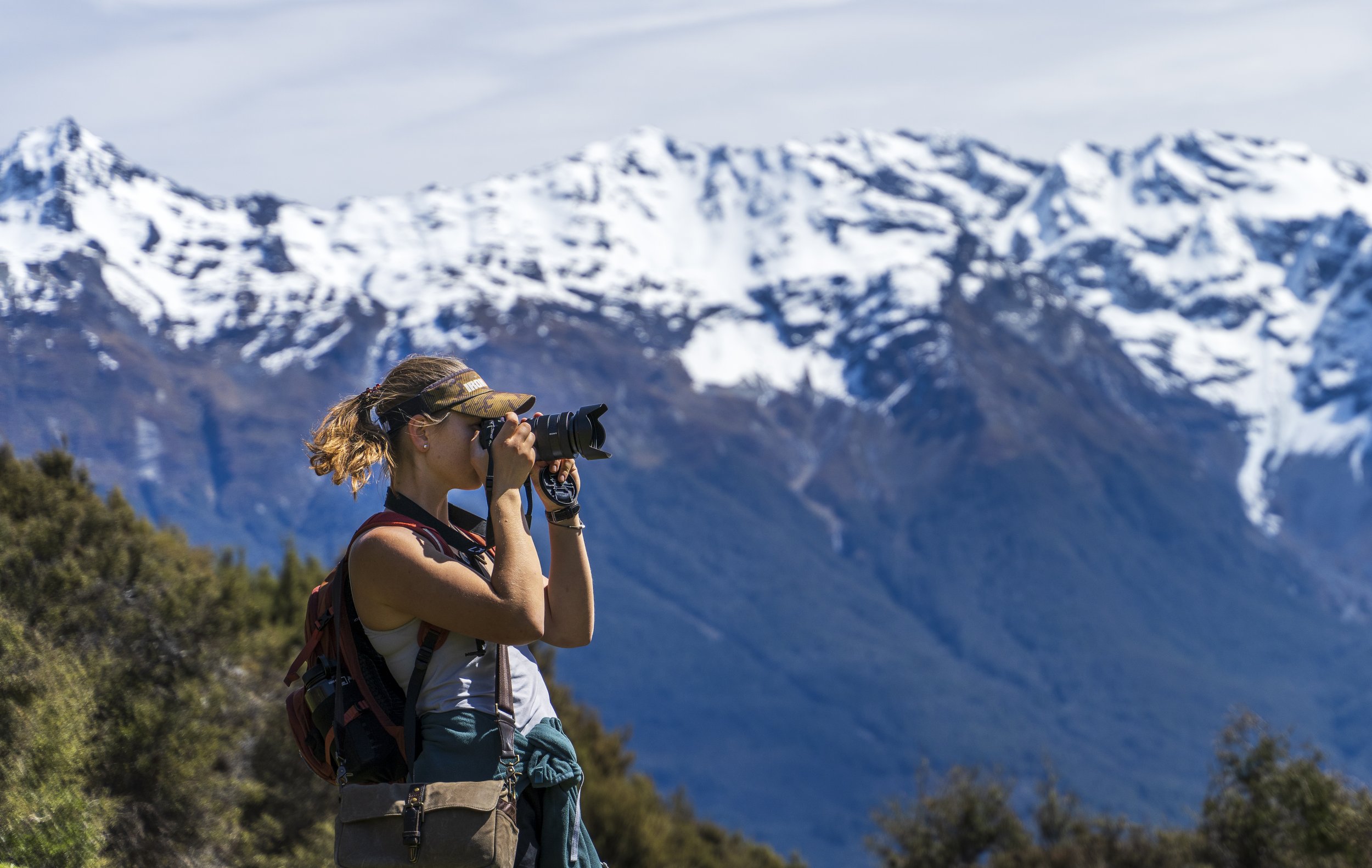 A woman standing outdoors with snow-capped mountains in the background, wearing a camouflage visor, taking a photograph with a camera.