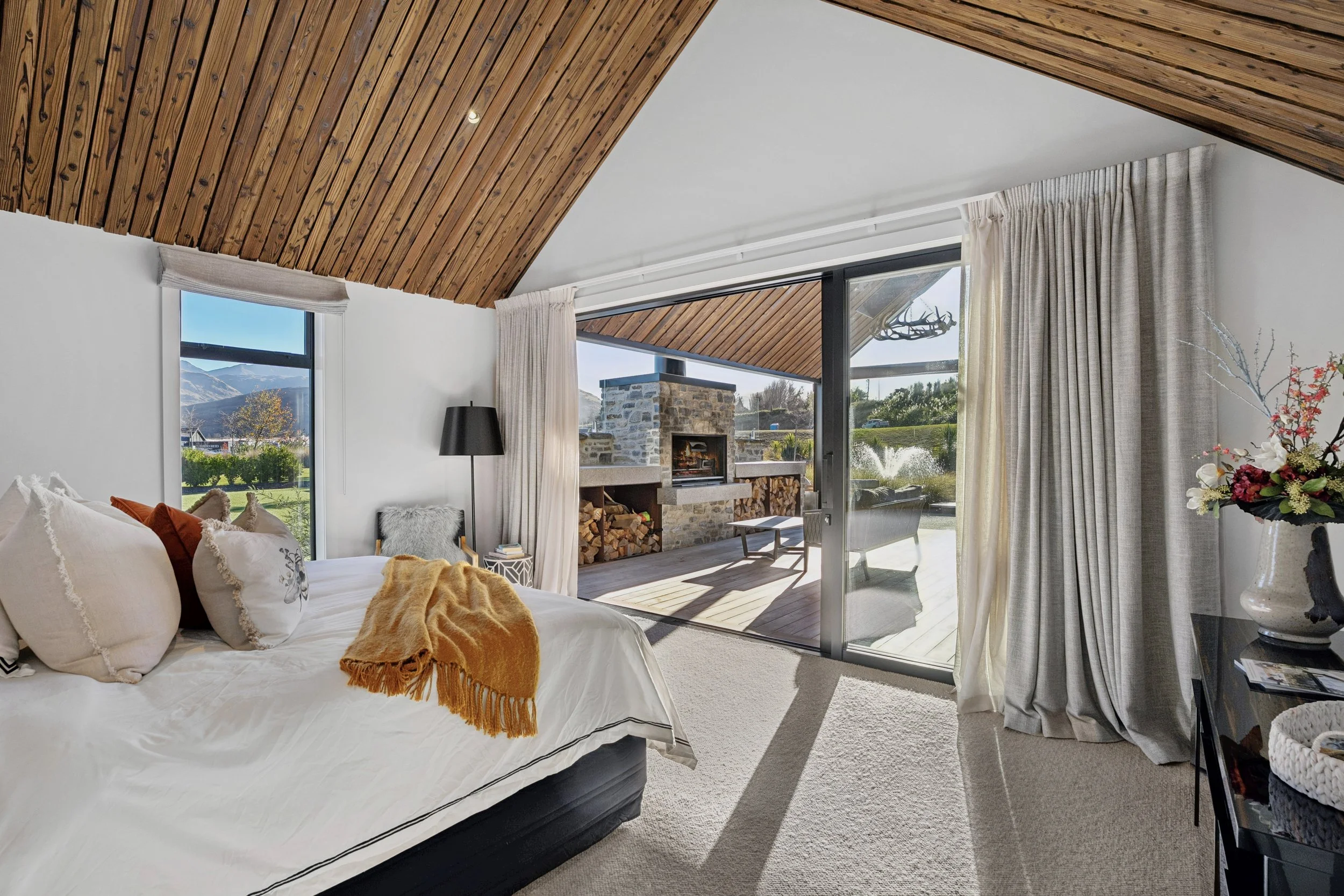 A cozy bedroom with a bed that has white bedding and decorative pillows, a mustard-colored throw blanket, and a black bed frame. The room features large windows with cream curtains, opening to a patio with a stone fireplace, wood storage, and outdoor