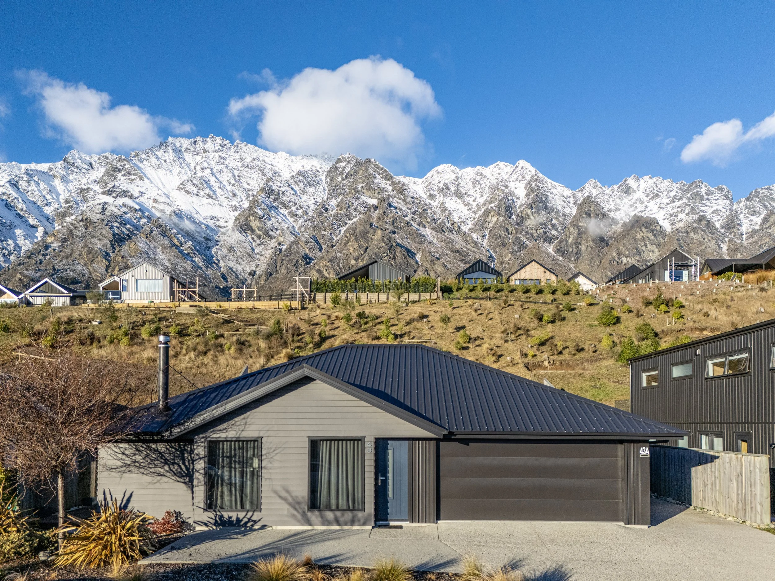 A residential house with a gray exterior and black roof in the foreground, with snow-capped mountains and a blue sky with clouds in the background.