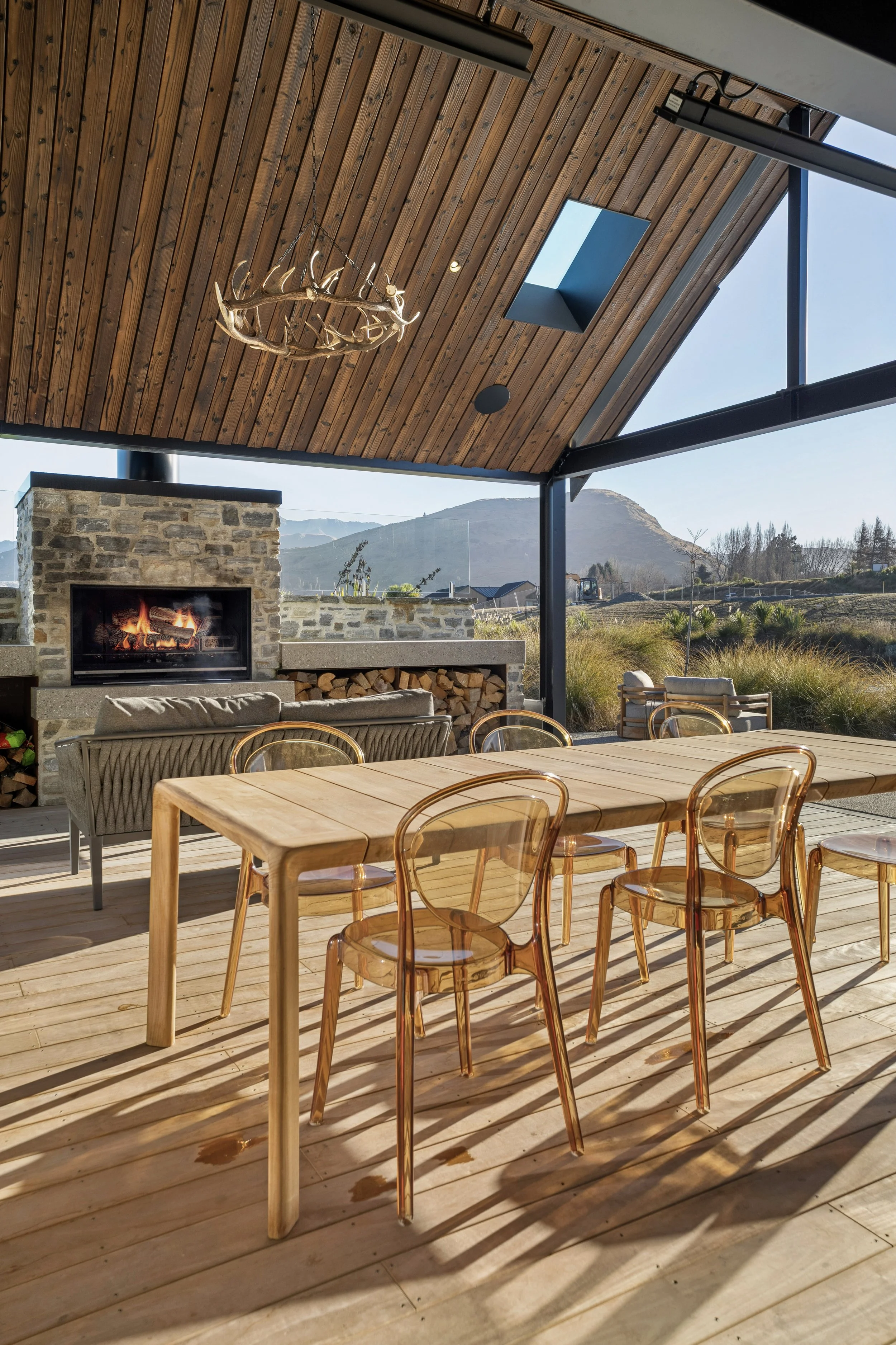 An outdoor patio area with a wooden ceiling, a stone fireplace, a large wooden dining table with transparent chairs, and a mountain landscape in the background.