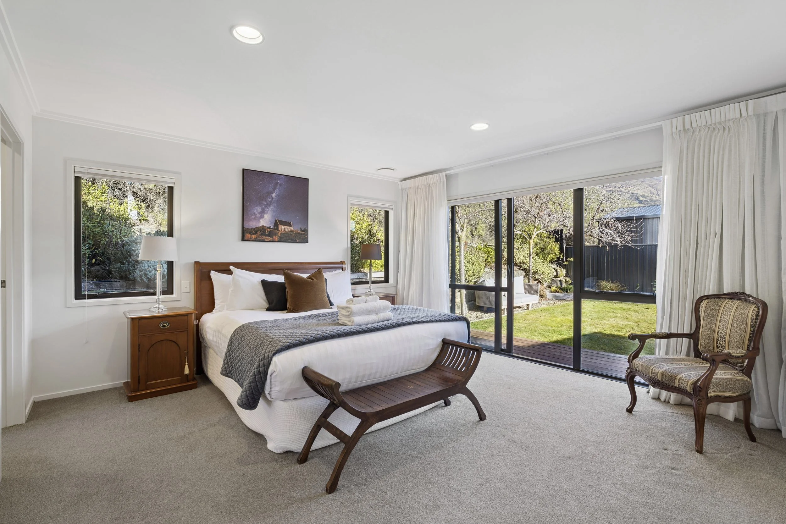 Comfortable bedroom with a large bed, wooden bench at foot, nightstands with lamps, and sliding glass door leading to outdoor patio and garden.