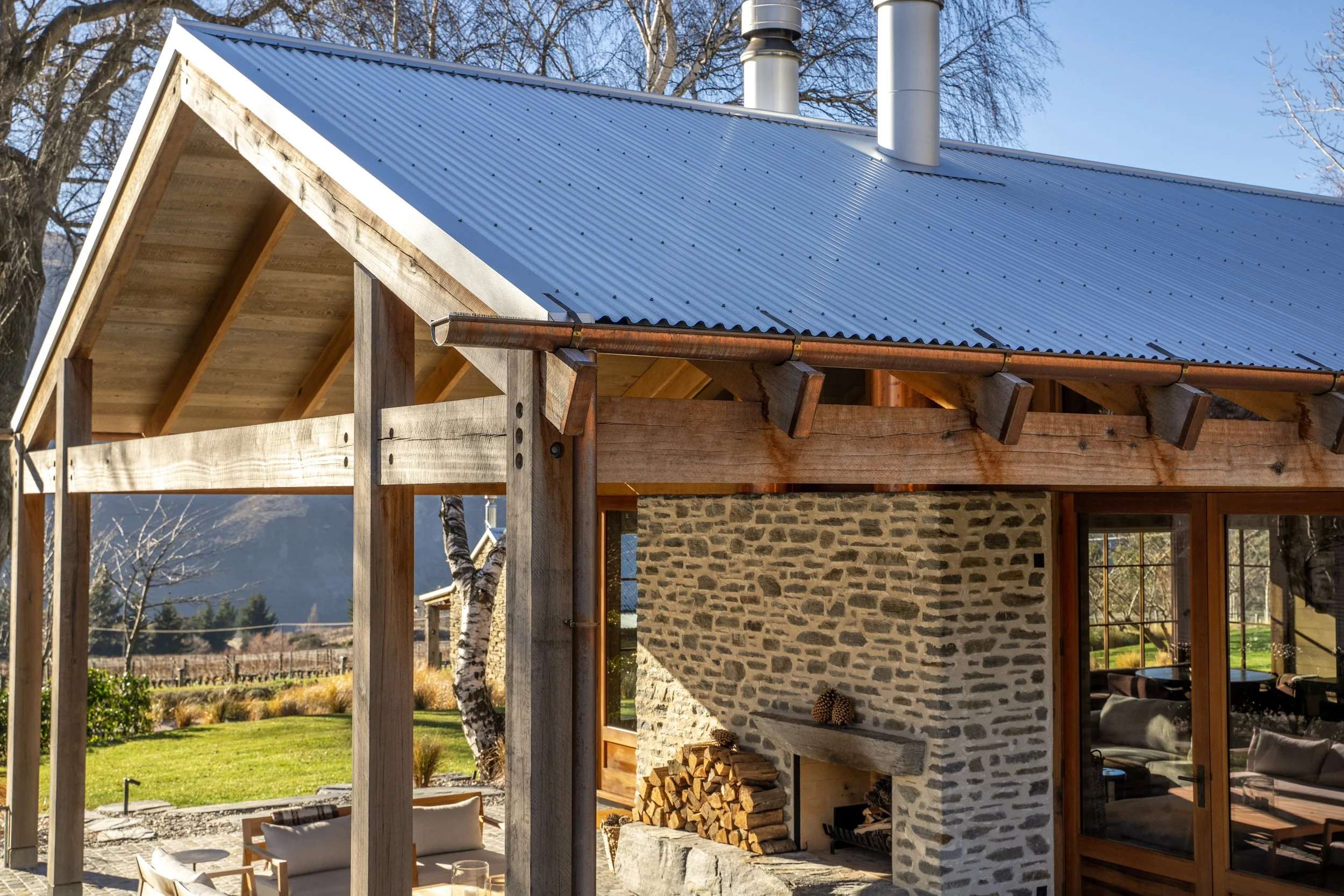 Partially constructed house with stone wall exterior, wooden beams supporting a corrugated metal roof, and an outdoor seating area with chairs and a stone fireplace showing wood logs inside.