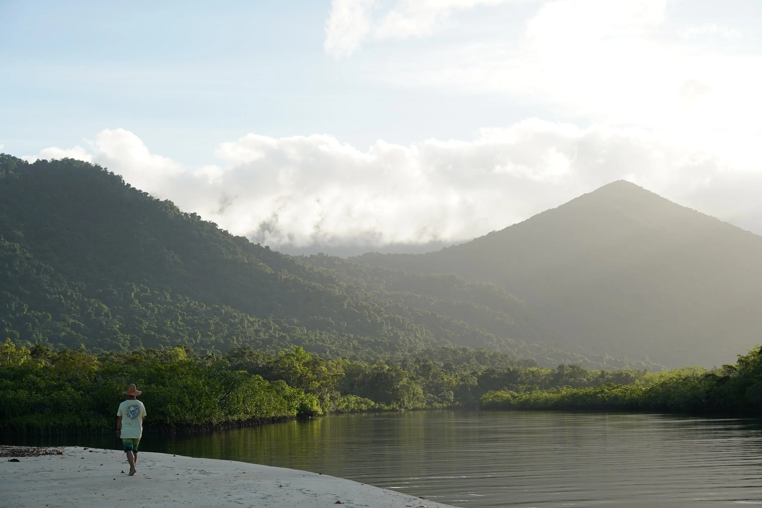 A person wearing a hat and white shirt walking along the sandy shore of a river, with lush green trees, mountains, and cloudy sky in the background.