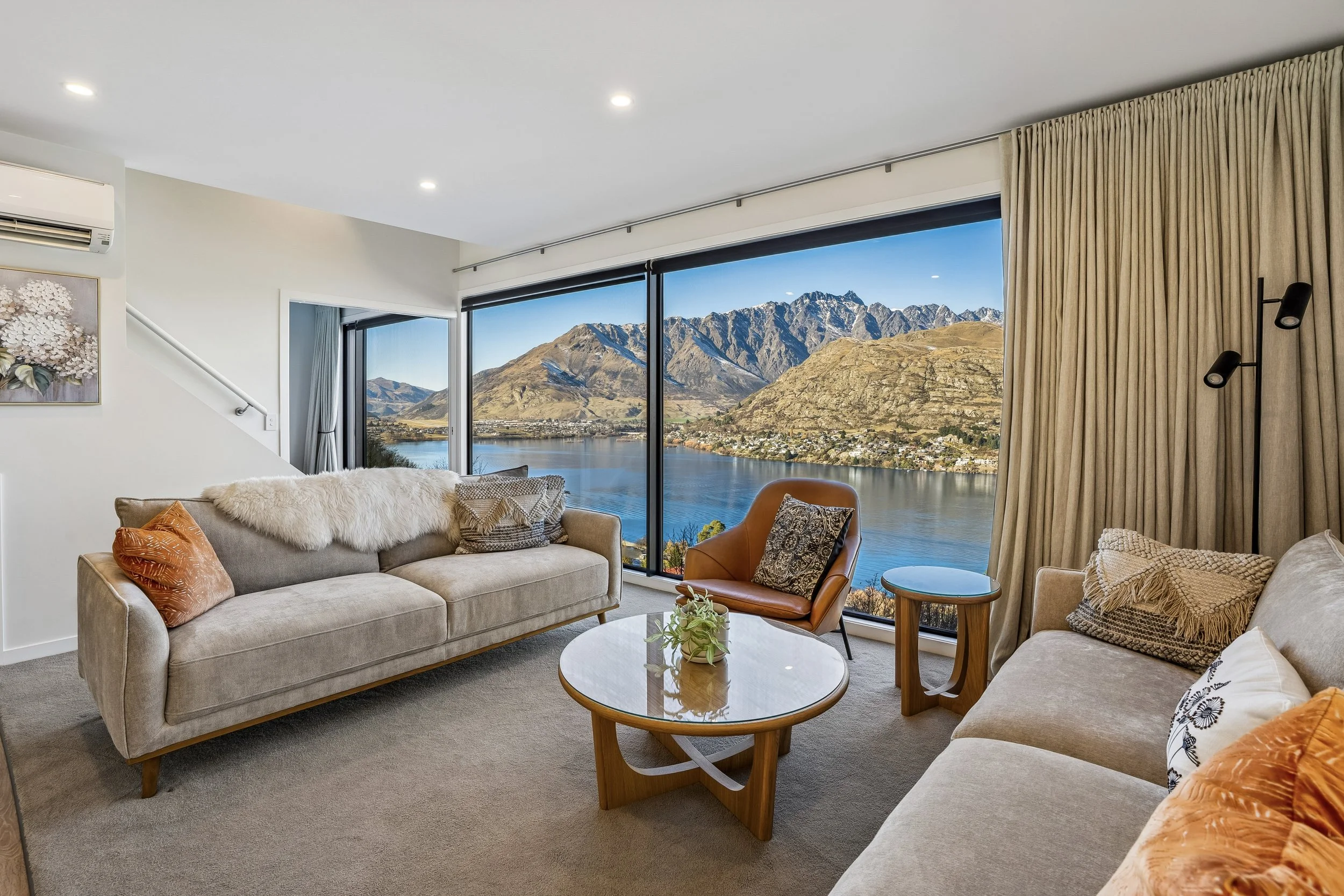Living room with large window view of the Queenstown mountains and lake Wakatipu, beige sofas with orange and patterned cushions, wooden coffee table with a small plant, and curtains on the side.