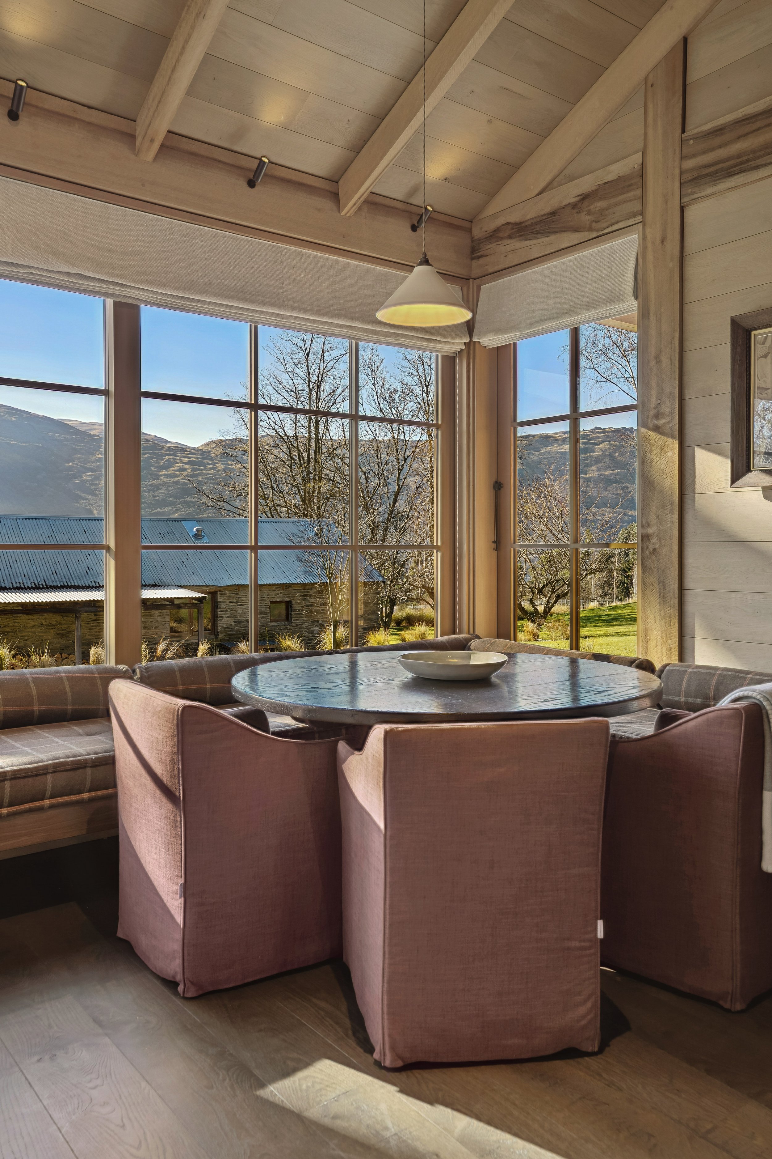 A cozy dining area with a round table surrounded by pink upholstered chairs, large windows showing a scenic mountain view, and wooden walls and ceiling, with sunlight streaming in.