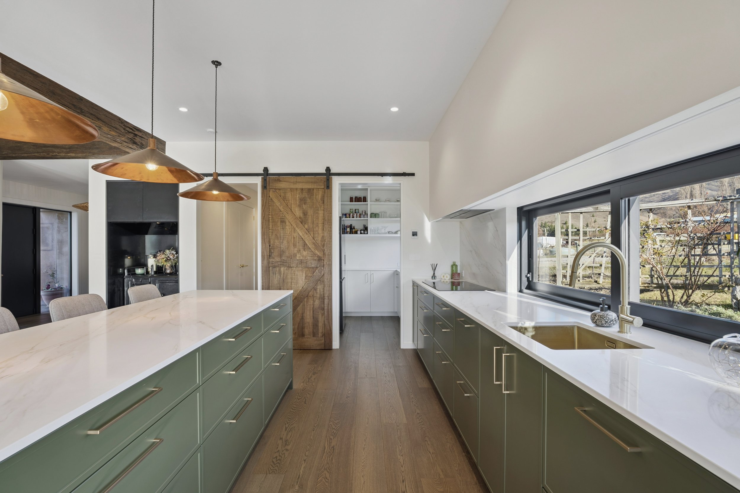 Modern kitchen with large window, green cabinets, white countertops, and gold fixtures.