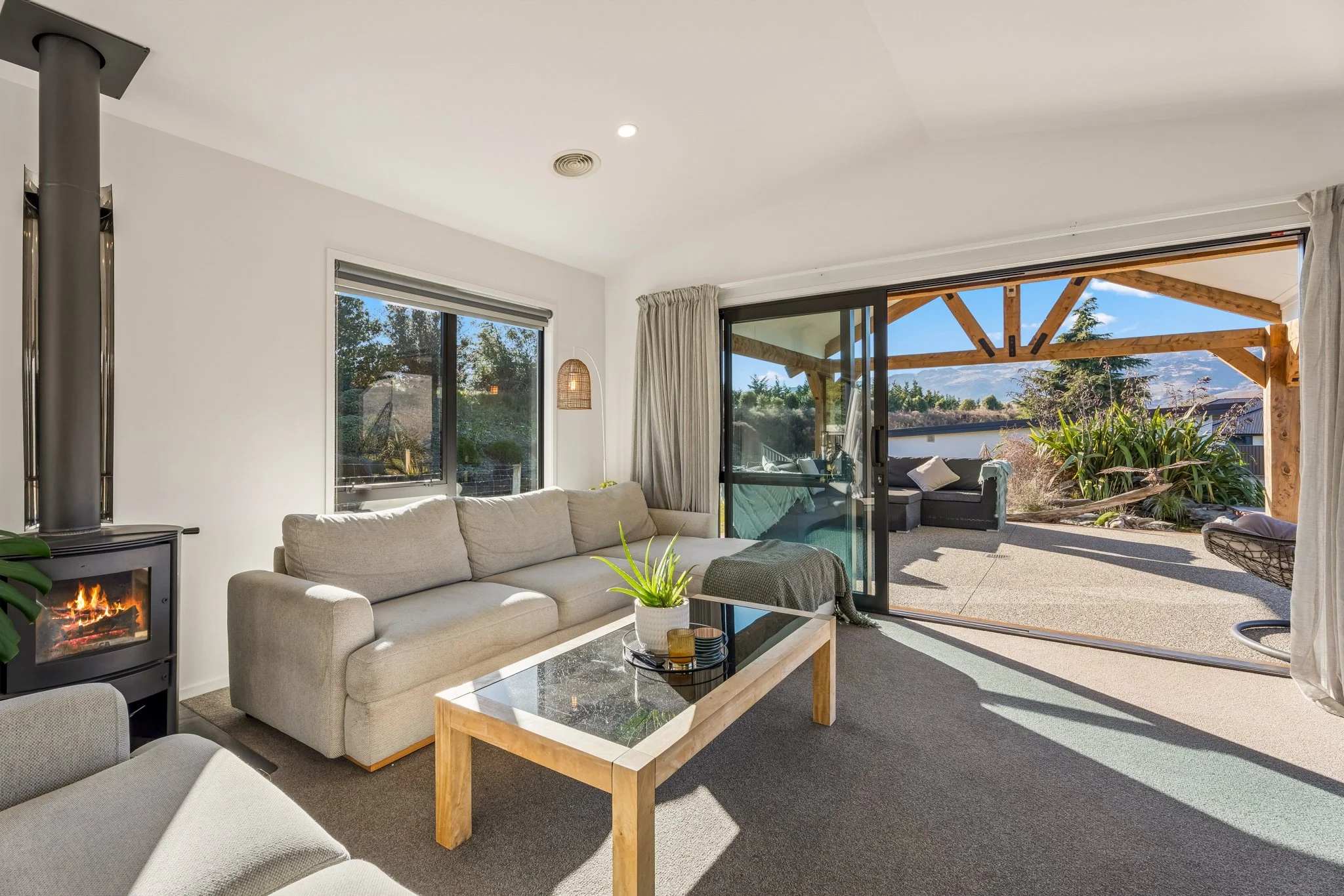 Living room with beige sofa, black fireplace, glass coffee table with plants, sliding glass door leading to patio with outdoor seating and wooden pergola, mountain view in distance.