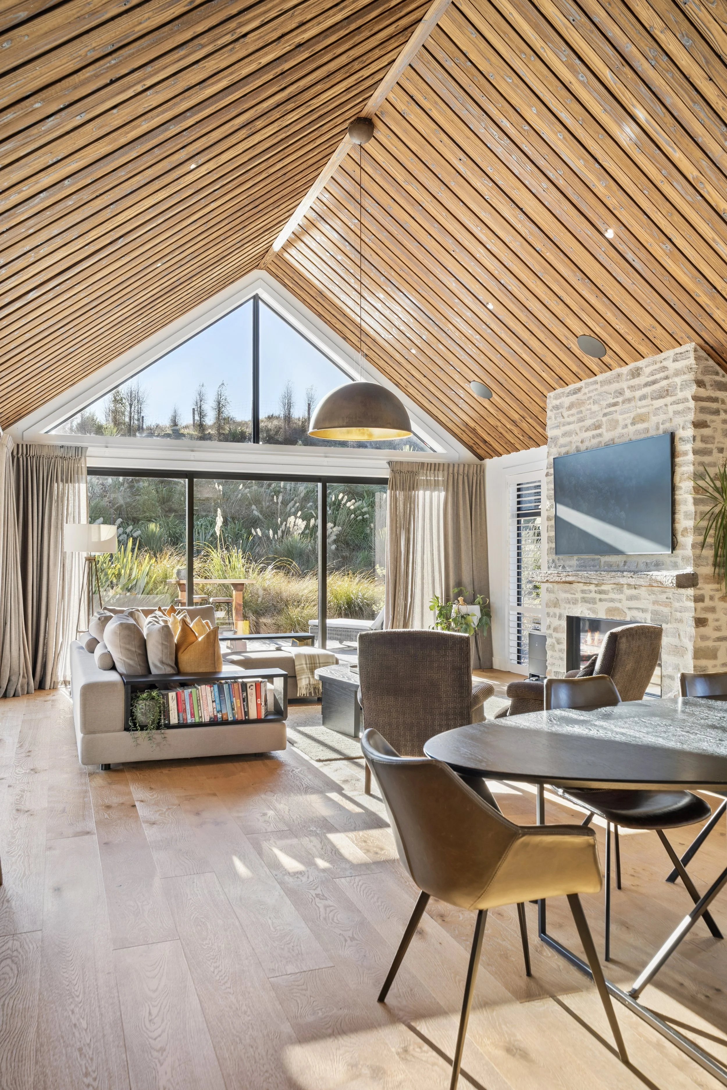 Modern living room with a vaulted wood ceiling, large windows, a stone fireplace with a TV mounted above, and various seating including a sofa, armchairs, and a dining table with chairs.