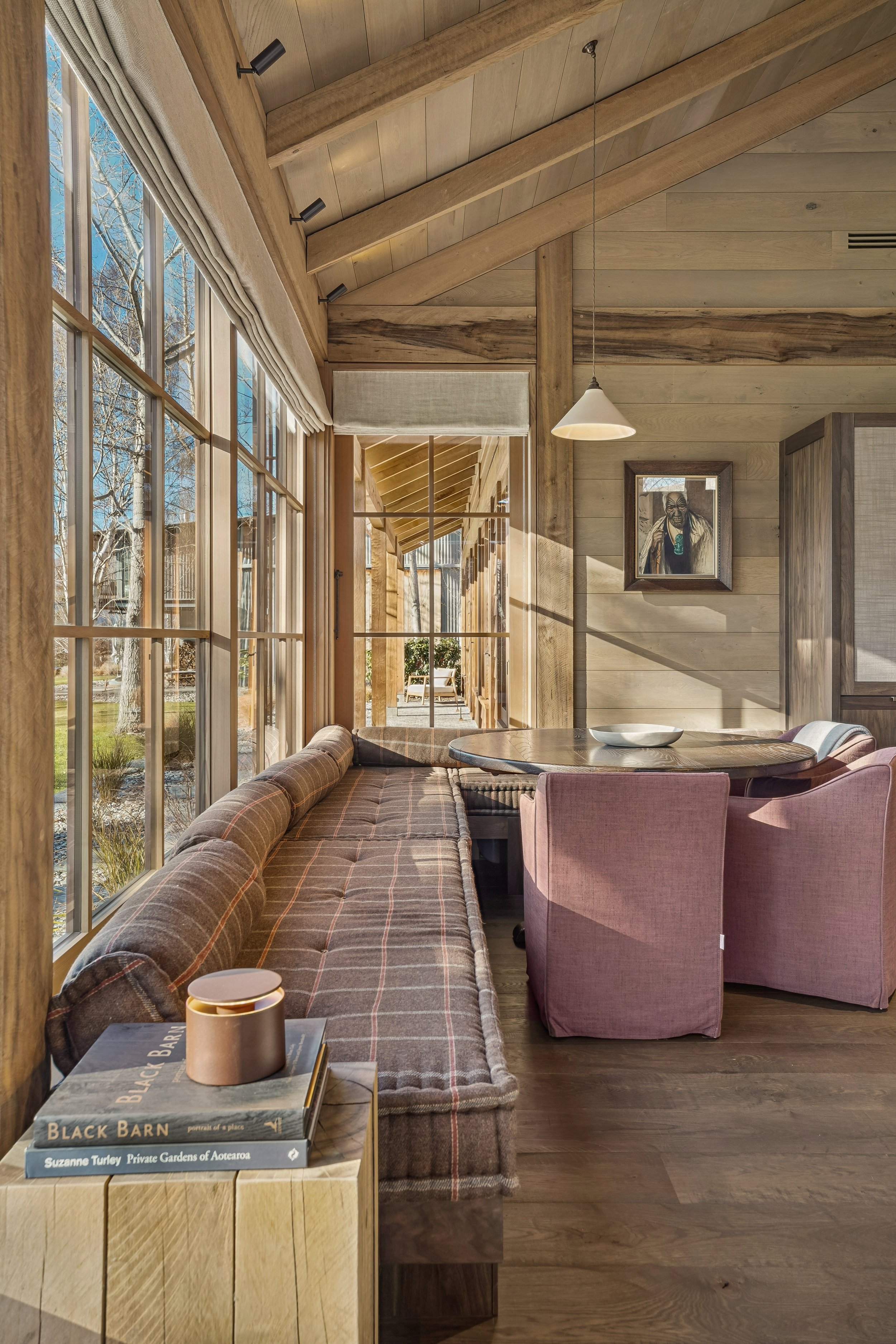 A cozy indoor space with wood paneling and large windows showing a sunny outdoor scene with trees. There's a round table with a pink upholstered chair and a plaid sofa along the window. A stack of books is on a wooden side table, with a candle on top