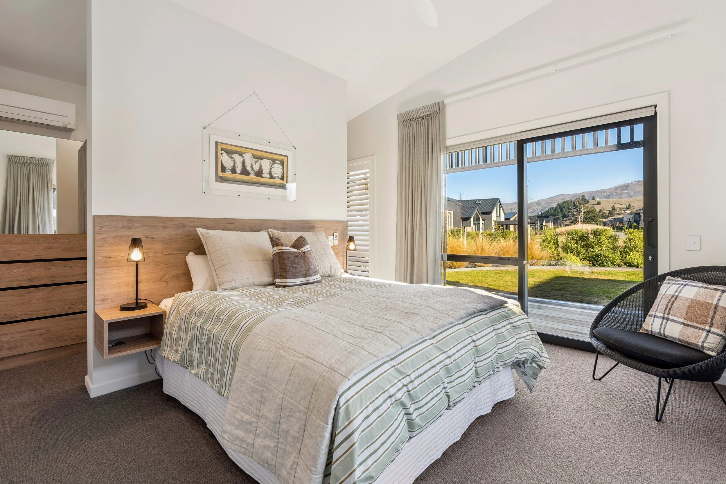 A bedroom with a large window showing a scenic outdoor view of mountains, houses, and greenery, a bed with plaid pillows, a wooden headboard, and a bedside lamp.