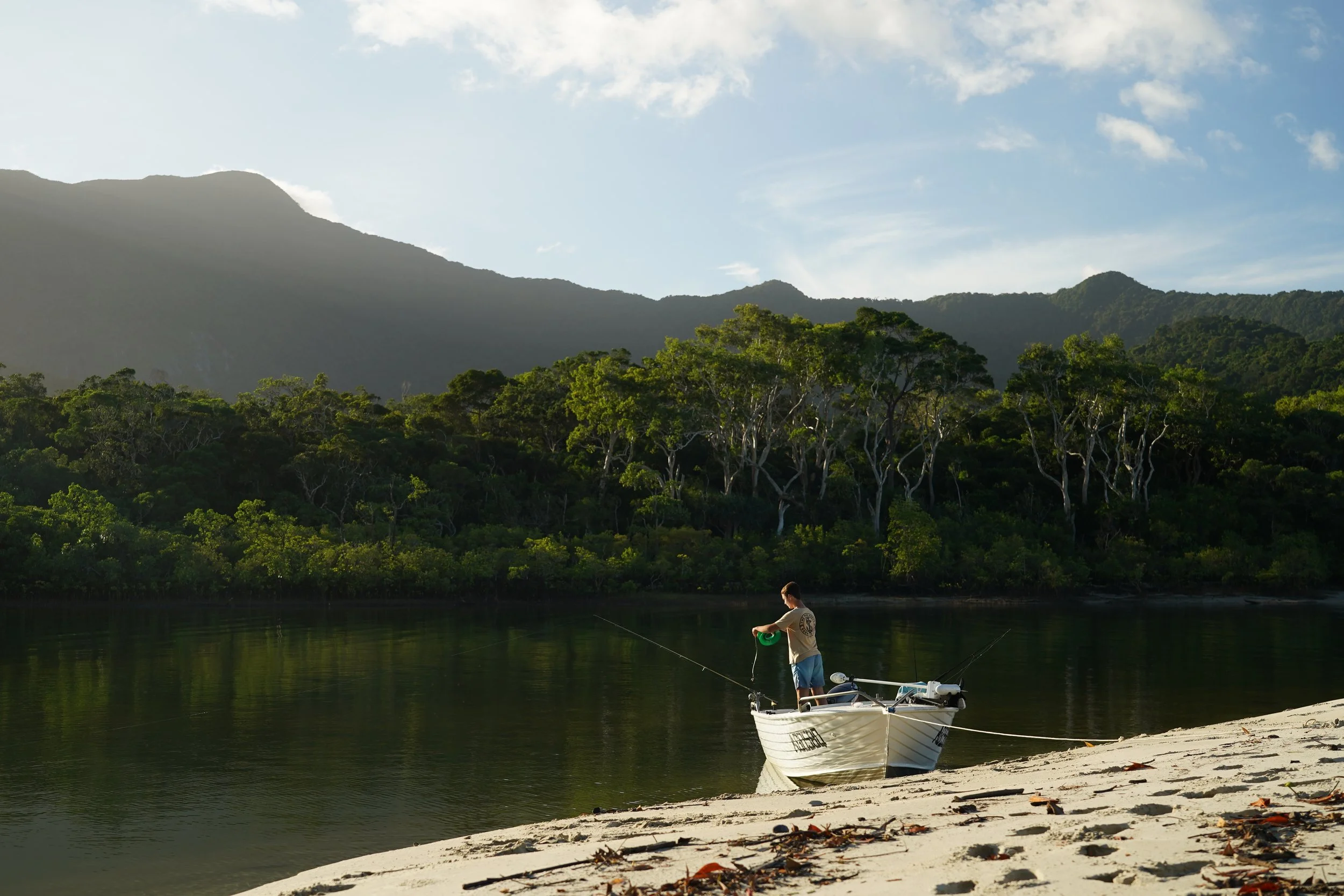 A person fishing from a small boat on a calm river near a sandy shore, with lush green trees and mountain hills in the background under a partly cloudy sky.