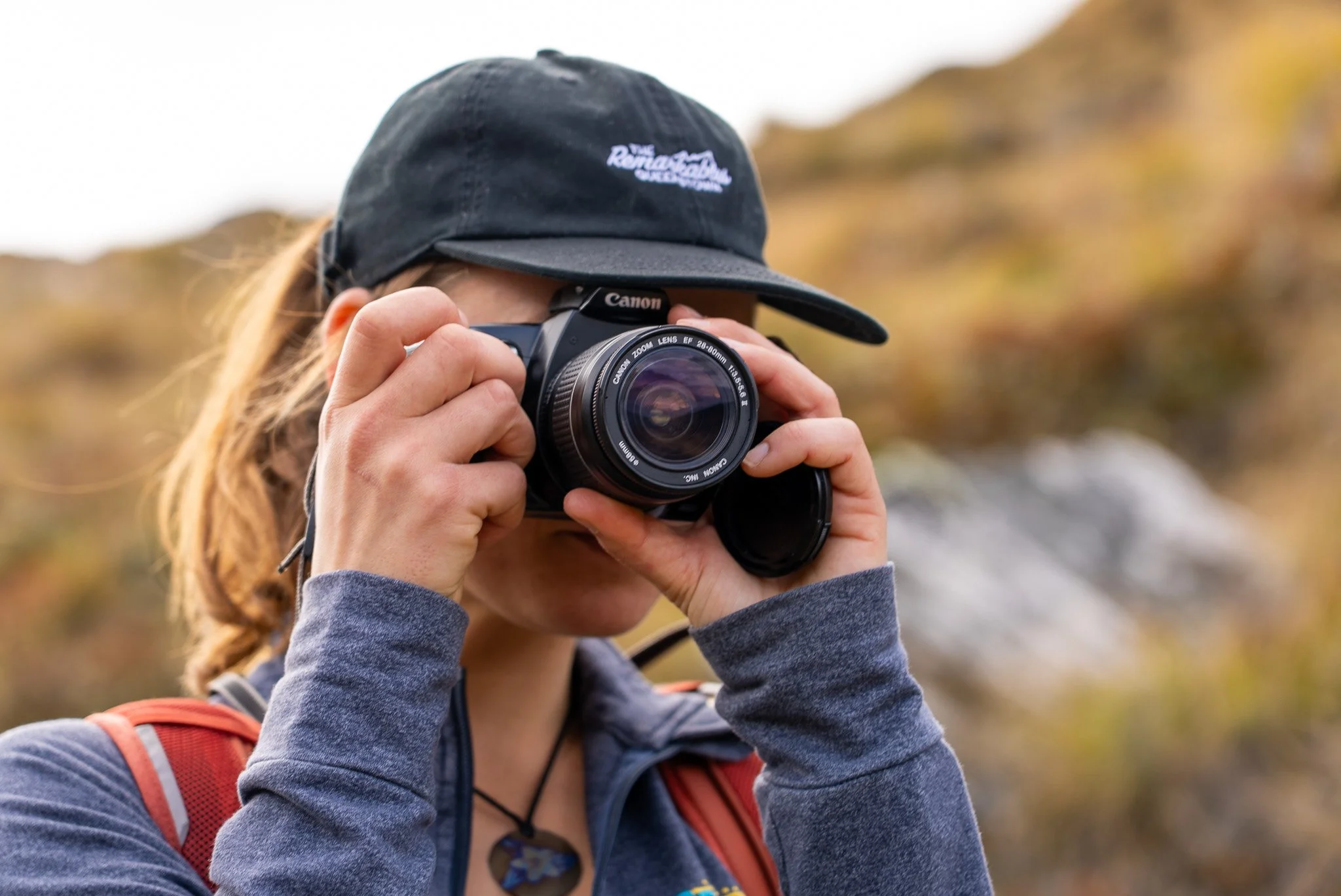 A person wearing a black cap and gray sweatshirt takes a photograph with a Canon camera outdoors, with blurred autumn foliage in the background.