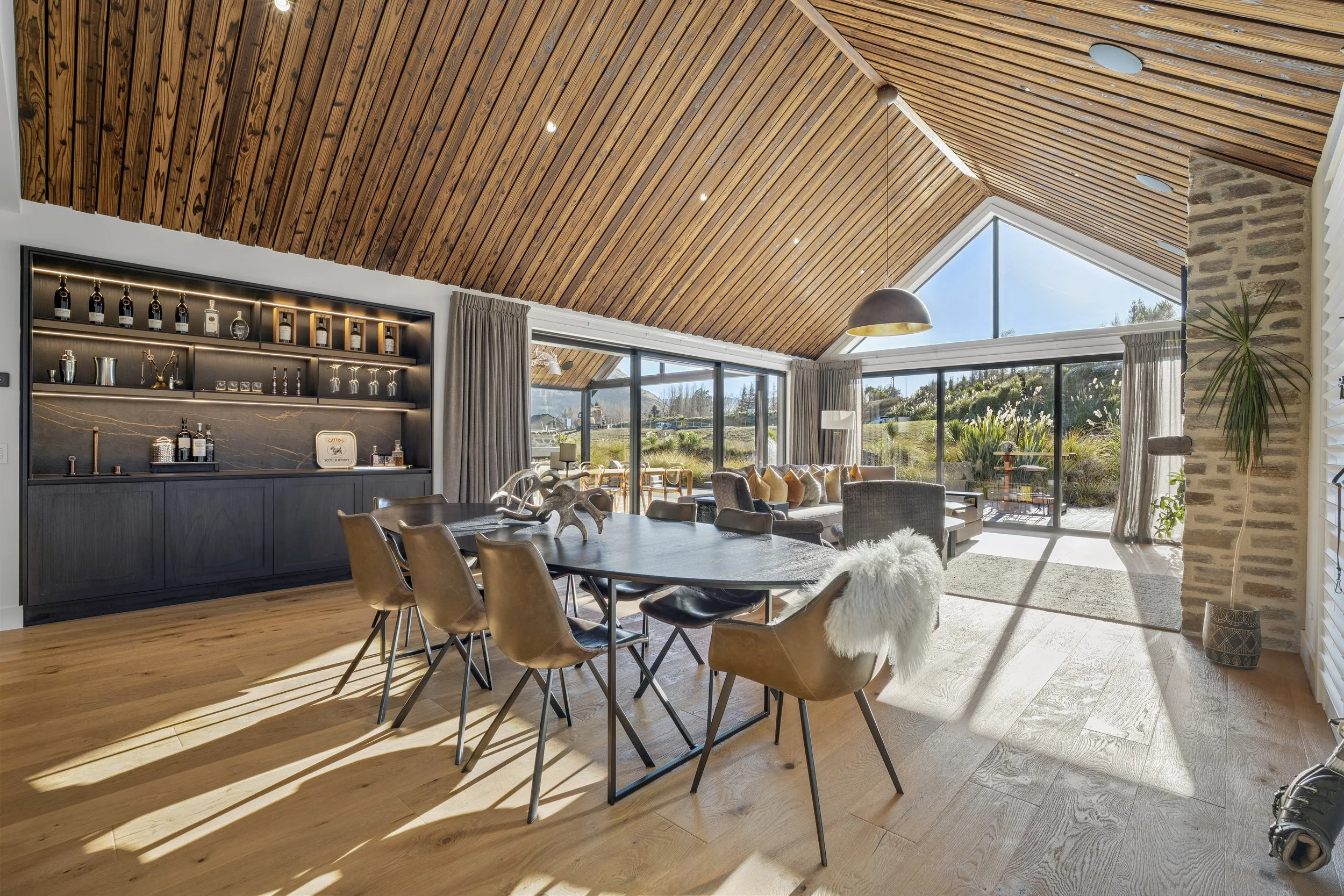 A spacious interior room with a high, sloped wooden ceiling and large windows showing a scenic outdoor view. The room features a dining table with chairs, a cozy sitting area with a sofa and armchairs, a built-in bar with bottles and glassware, and v