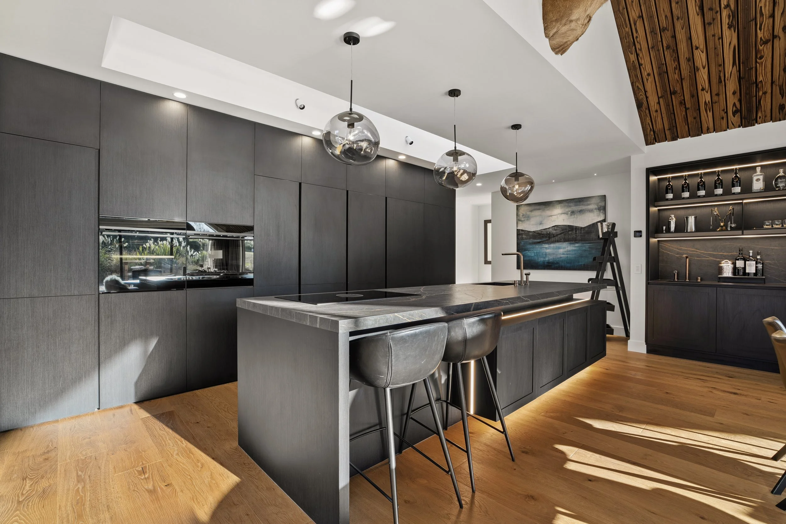 Modern kitchen with dark cabinetry, a marble island with barstools, pendant lights, and wall-mounted shelves with bottles and glassware.