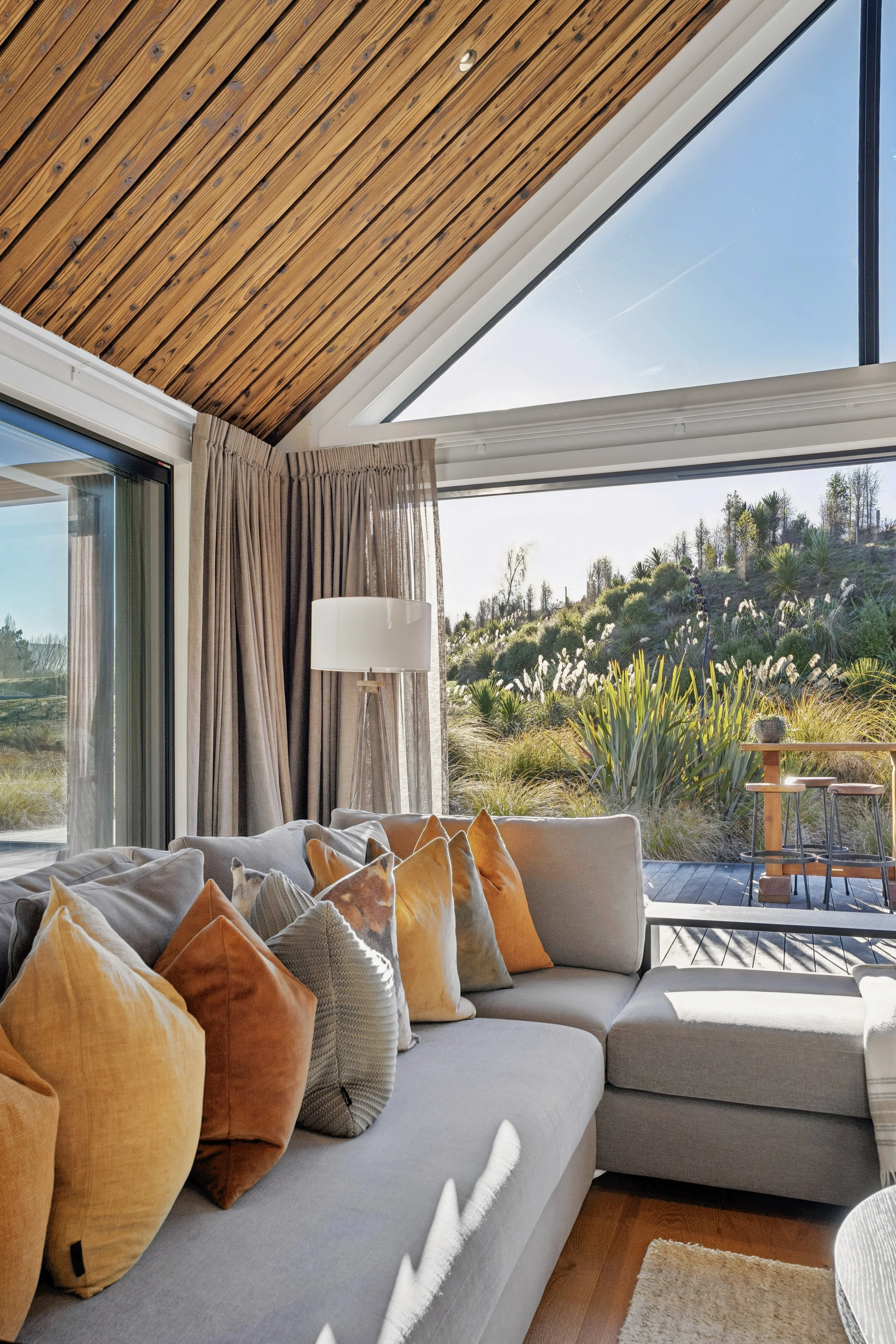 A modern living room with a large L-shaped gray sofa decorated with orange, beige, and patterned pillows. The room has large floor-to-ceiling windows revealing a scenic outdoor view of plants and trees. The ceiling is made of wood planks, and there a