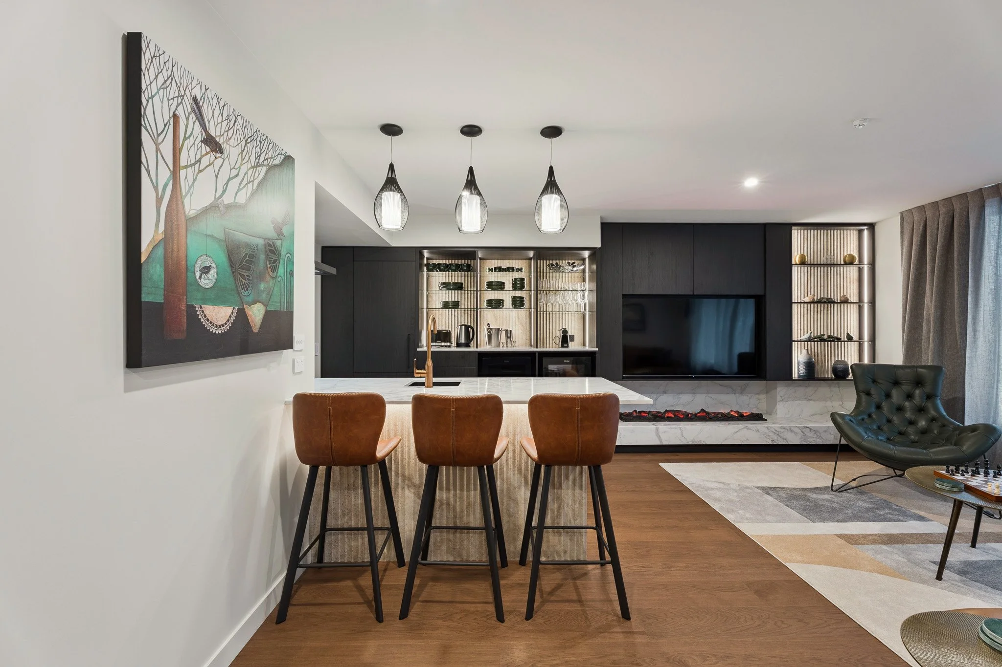 Modern kitchen and living space with a table, three brown chairs, black pendant lights, black cabinetry, built-in oven, glass shelf with dishes, a large TV, a corner shelf with decorative items, and a black and gray lounge chair.