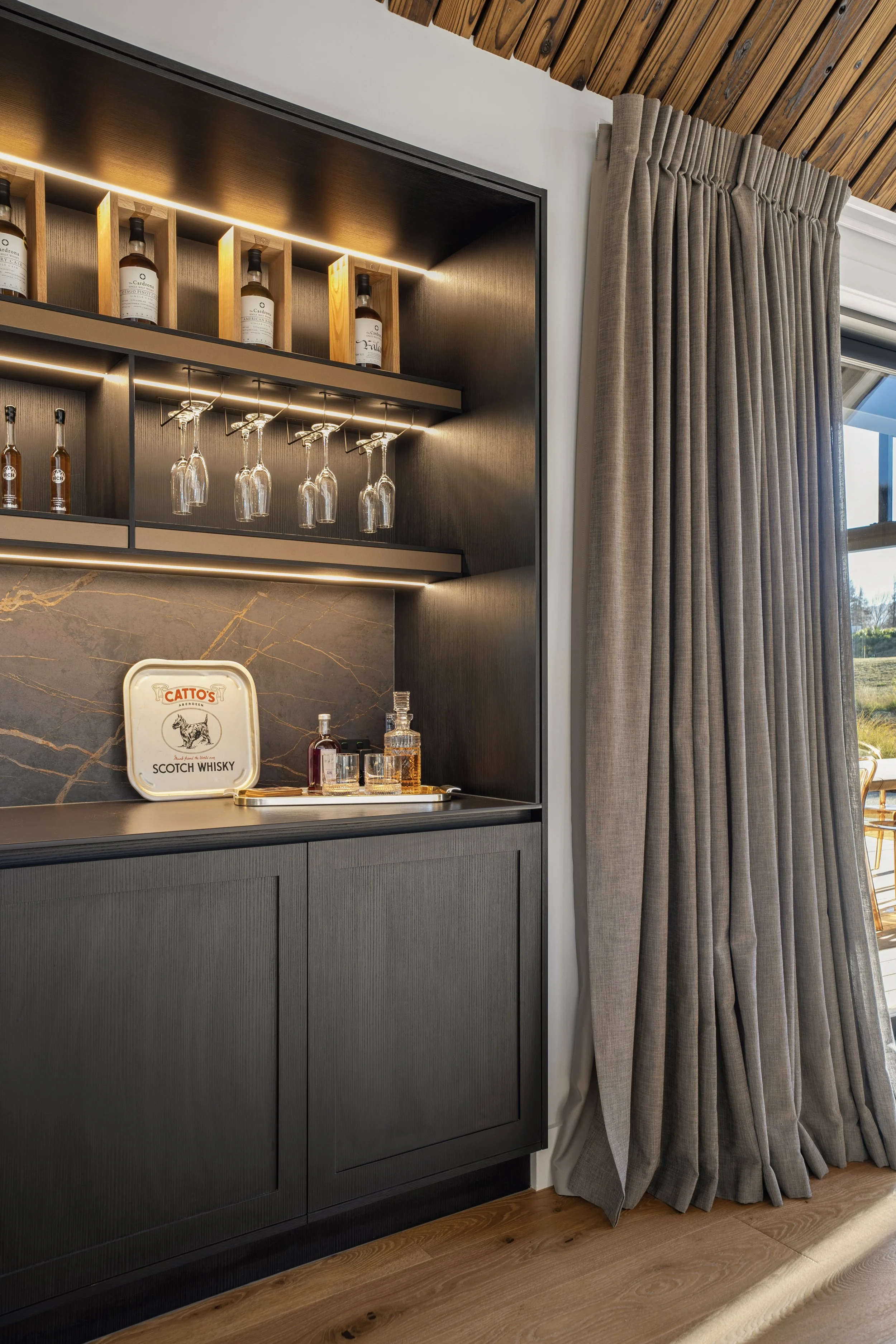 Close-up of a modern kitchen bar area with dark gray cabinetry, a marble backsplash, and a tray with glasses and bottles of whiskey. Behind it, a decorative shelf displays liquor bottles, and under the shelf, wine glasses hang upside down. A large wi