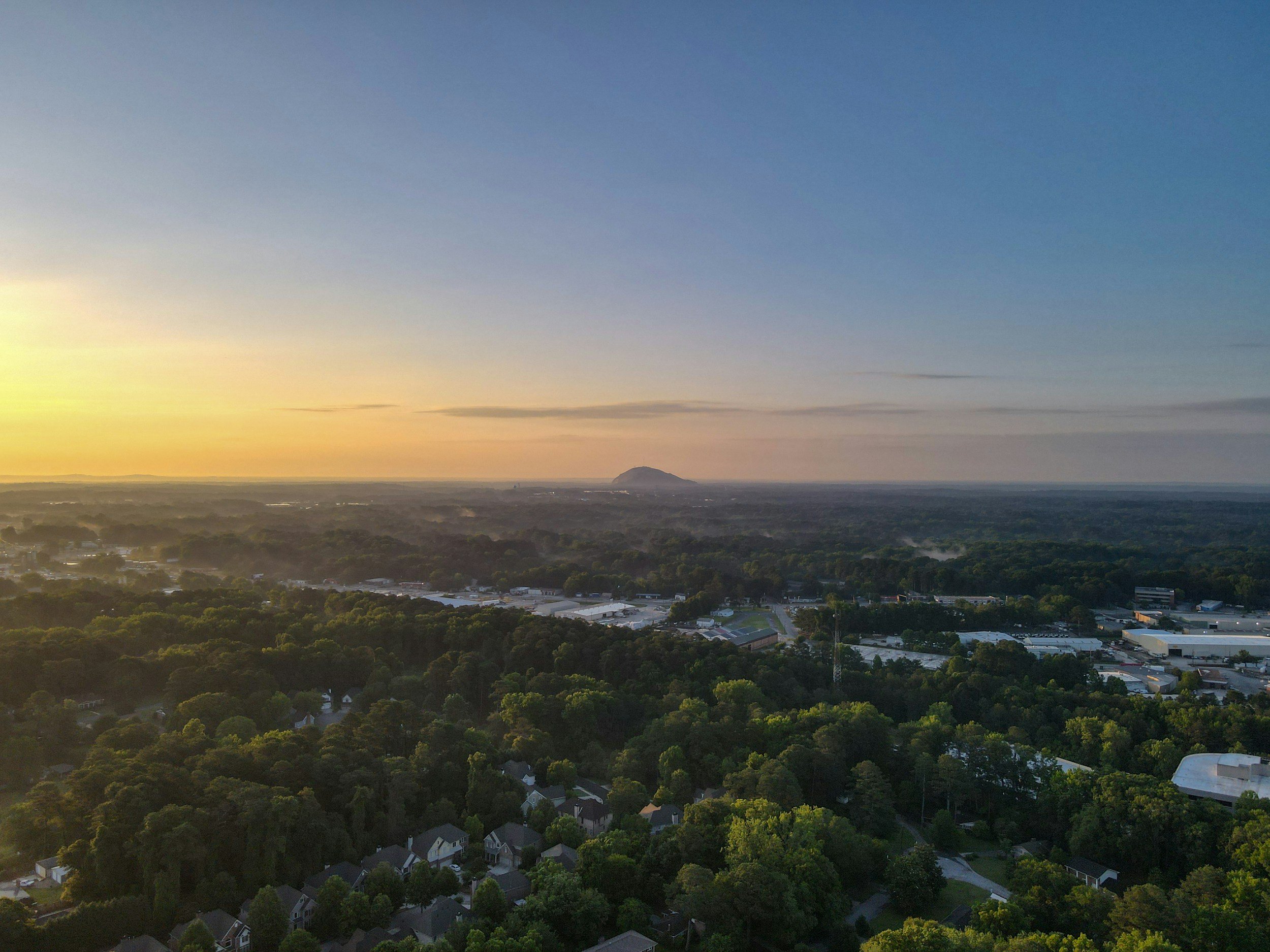 Aerial view of a suburban area with trees, houses, and industrial buildings during sunset, with a mountain in the distance and a clear sky.