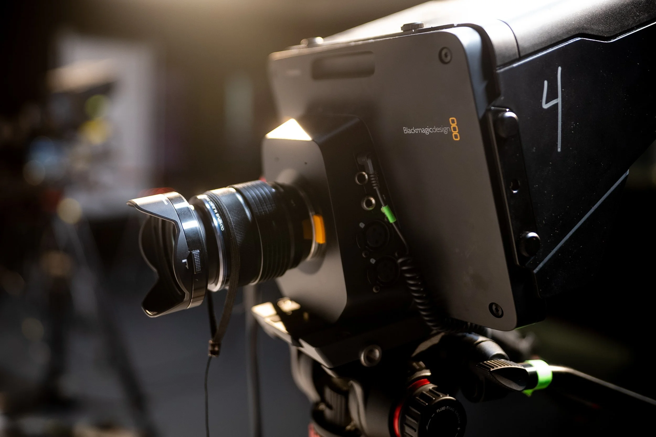Close-up of a professional video camera with a large lens and connectors, mounted on a tripod, in a dimly lit studio. The camera is labeled '4' and features controls and a microphone input.