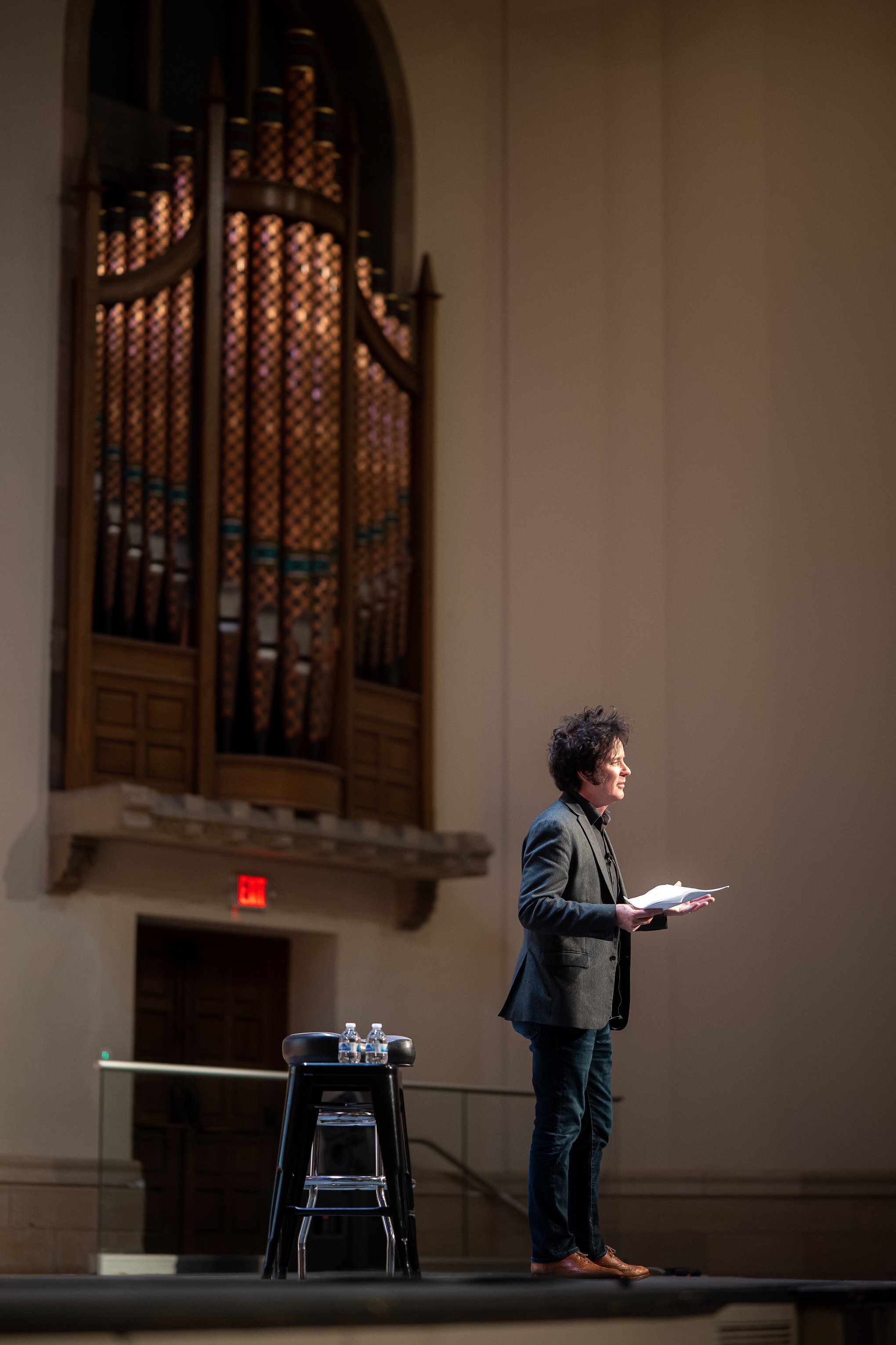 A person speaking on stage in a large venue, with a pipe organ in the background and a stool with water bottles beside them.